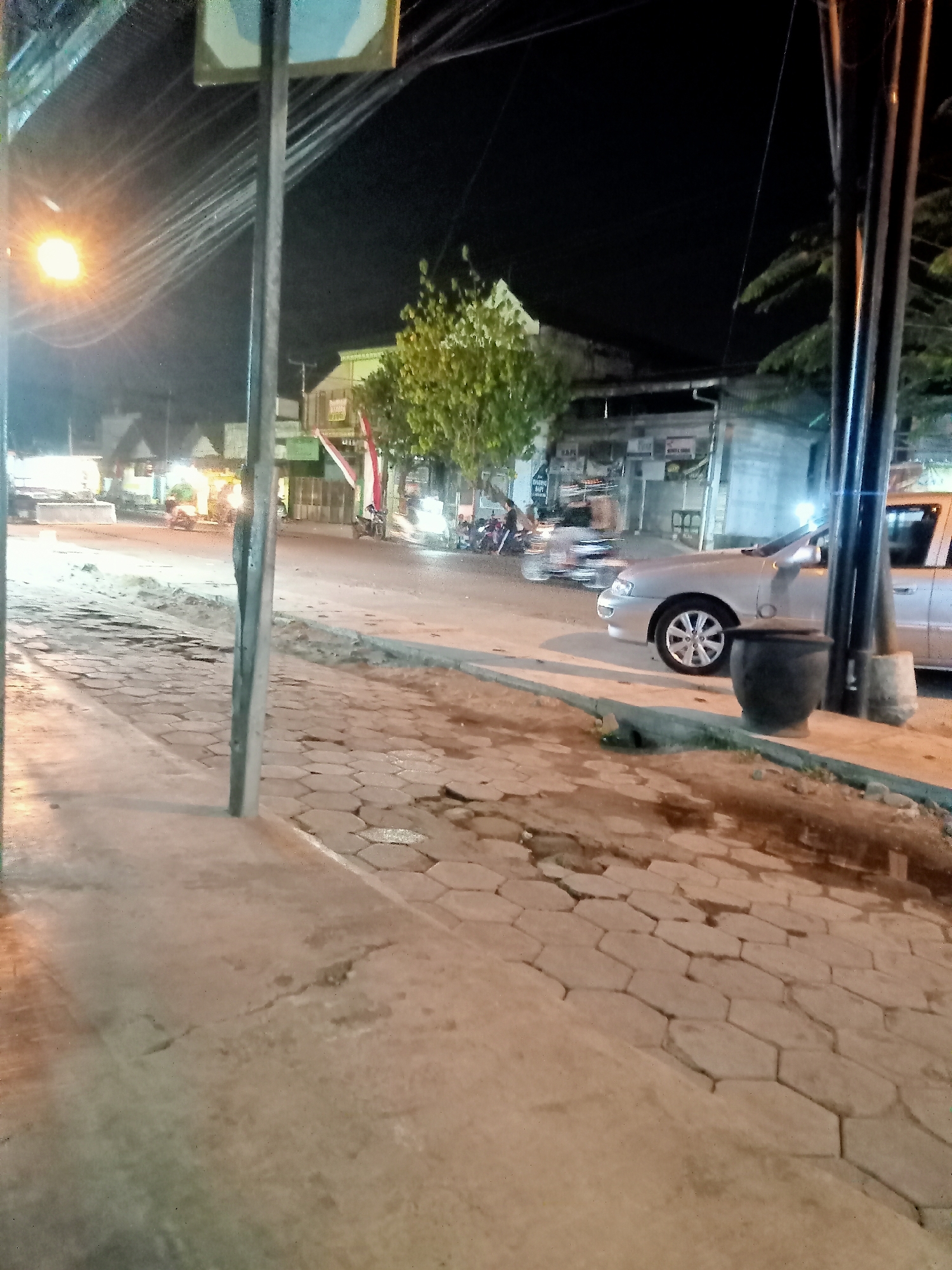 The image captures a dimly lit street scene at night, likely in Mangunsari, Indonesia, evidenced by the presence of street lighting and typical Indonesian urban infrastructure. A silver sedan is parked on the right side of the road, with a large black trash bin positioned in front of it. The foreground features a cracked and uneven paved sidewalk with hexagonal stones, suggesting wear and tear. Further down the street, several motorcycles are visible, some with riders, and various shops and buildings line the road, illuminated by artificial light. A street lamp with a warm glow is prominent on the left side of the frame, casting long shadows and contributing to the overall nocturnal ambiance. The sky is dark, indicating nighttime.