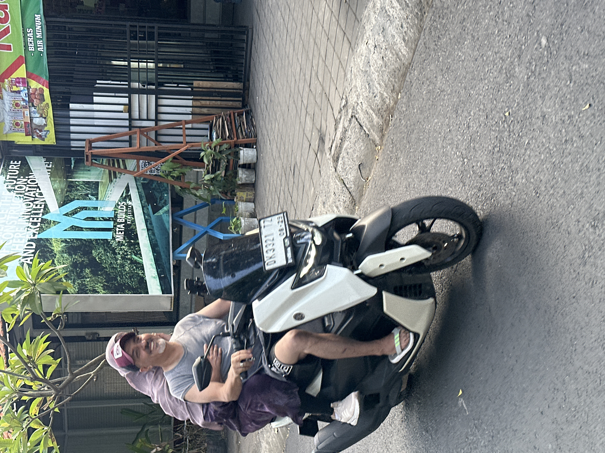 In North Kuta, Indonesia, a man and a woman ride a white and black scooter on a paved road. The man is driving, wearing a gray t-shirt, black shorts, a maroon baseball cap, and a ring on his left hand. He has a mustache and goatee. The woman is behind him, wearing a purple headscarf and a dark purple garment. Her left foot is visible, wearing a white sneaker with green and grey accents. The scooter has a black windshield and a license plate reading "DK 3321 TZ". In the background, a building with an advertisement for "META BUILDS REAL ESTATE" is visible, along with potted plants, a ladder, and a metal fence. The ground is a mix of asphalt and grey paving stones.