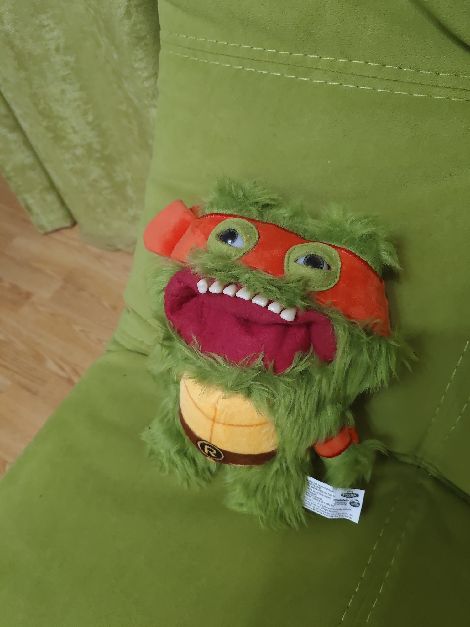A plush toy, resembling the Teenage Mutant Ninja Turtle character Michelangelo, is positioned on a green, textured sofa. The toy is predominantly green and furry, with orange accents around its eyes and a red bandana. It has a wide mouth with visible white teeth and a red tongue. A yellow plastron with a brown trim and a red "R" insignia is visible on its torso. The sofa has decorative stitching. The background shows a wooden floor and another part of the green sofa. A white tag is attached to the toy's arm. The lighting suggests an indoor setting. There is no discernible indication of the city of Rostov-on-Don, Russia, within the image itself.