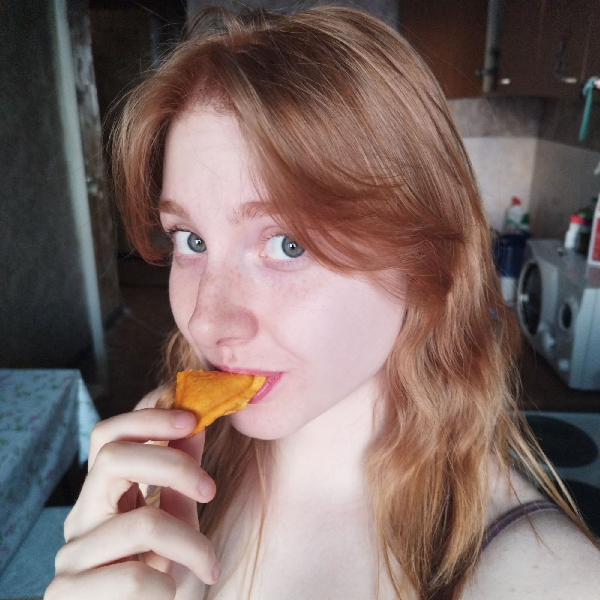A close-up, eye-level portrait shows a young woman with fair skin, freckles, and long, wavy reddish-brown hair. Her light blue-grey eyes are wide open and look directly at the camera. She has full, pink lips, slightly parted, with a piece of orange-yellow food, possibly a fried dumpling or pastry, held up to them between her thumb and index finger. A dark, possibly brown, strap is visible on her right shoulder.
The background suggests an indoor home setting, likely a kitchen or dining area in Yekaterinburg, Russia. To her left, part of a table covered with a light-colored tablecloth, possibly with a faint floral pattern, is visible. Behind her, a kitchen environment is blurry but discernible, featuring dark-colored upper cabinets and a lighter countertop. Various household items are visible on the counter, including a blue container, bottles with white, red, and green labels (resembling cleaning products), and a white kitchen appliance (possibly a microwave or oven). A black circular shape, possibly a stove burner, is also vaguely visible in the far right background. The lighting is soft and even, suggesting an indoor setting with either diffused natural light or artificial lighting. Her expression appears candid and somewhat inquisitive.