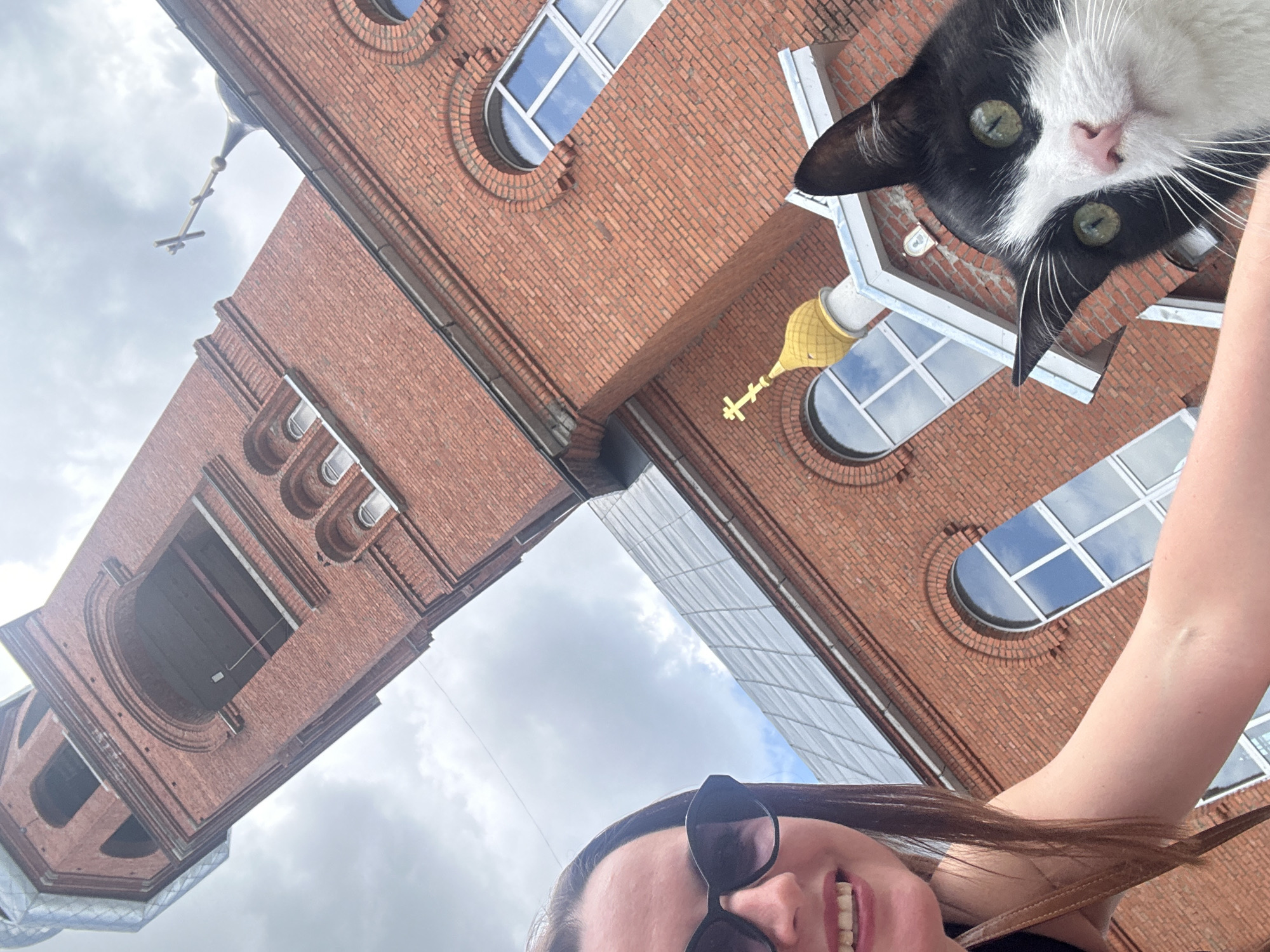 A low-angle, selfie-style photograph captures a woman and a black and white cat in front of a red brick church in Medvedevo, Russia. The woman, wearing sunglasses and a black top, is on the left side of the frame, her arm reaching out. Her mouth is open in a smile. To the right of the woman, a black and white cat with green eyes is prominently featured, looking directly at the camera. The cat's ears are perked up. Behind them, the tall, red brick bell tower of the church dominates the background. Architectural details of the church, including arched windows and a golden dome with a cross, are visible. The sky is cloudy.
