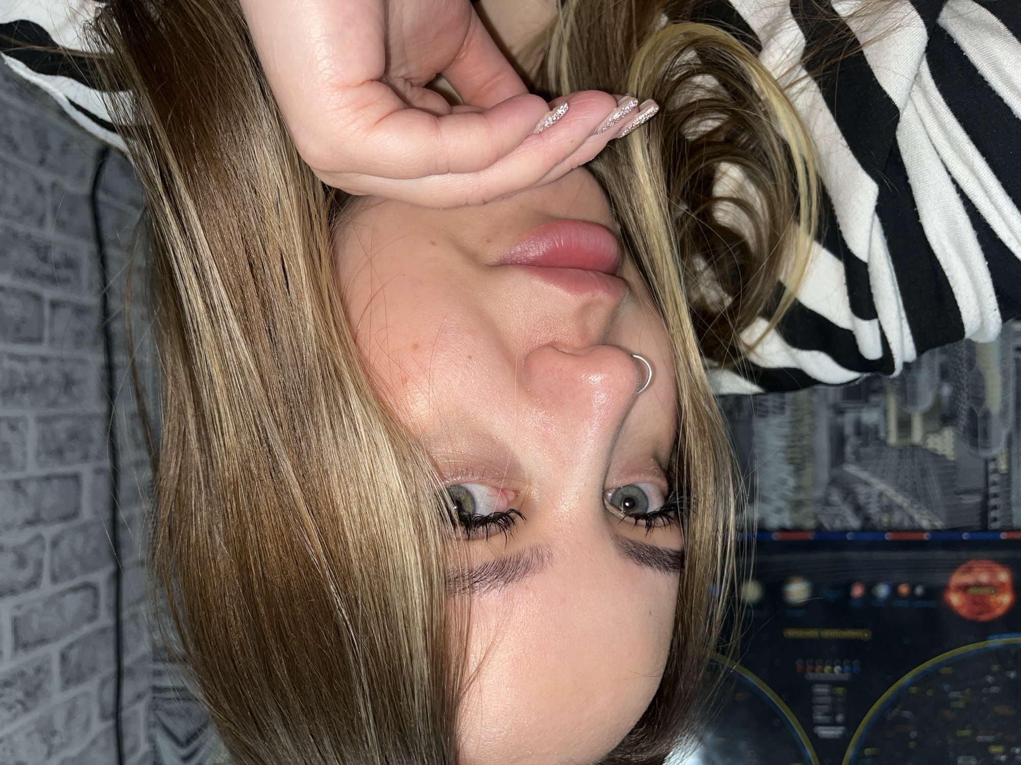 A woman with blonde hair, a nose ring, and decorative fingernails poses indoors. She is wearing a black and white striped top. In the background is a wall with a map of the solar system on it. There is also a wallpaper with a brick pattern. There is no indication that the image was taken in Novopokrovskaya, Russia.