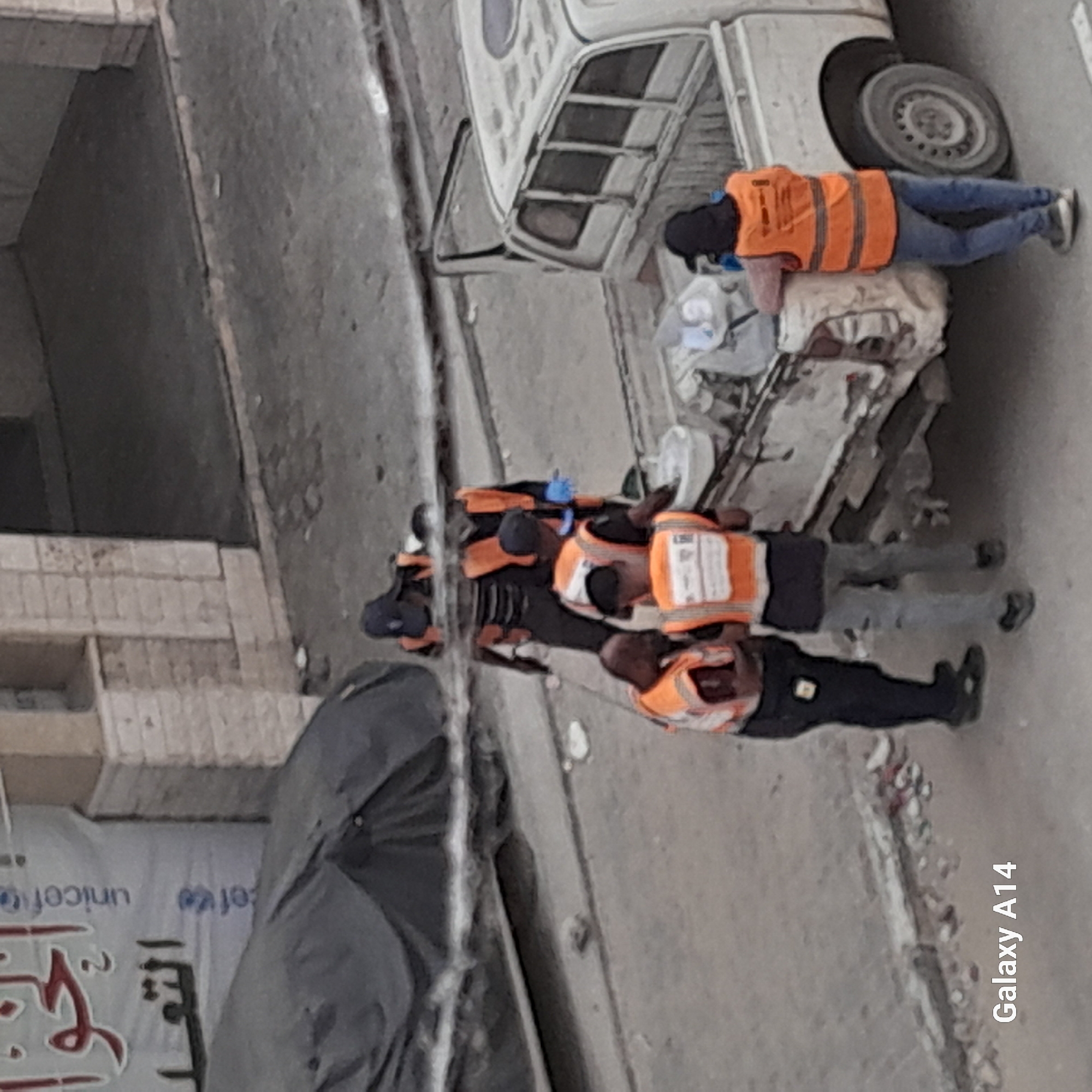 In Khan Yunis, Palestinian Territories, a group of individuals wearing orange safety vests are gathered near a white pickup truck. The scene appears to be outdoors, with a paved surface in the foreground and a building with textured walls in the background. Some individuals are looking towards a white bucket or container. One person is leaning against the back of the pickup truck, which has its rear door open and is loaded with various items. The overall impression is of a work or cleanup operation.