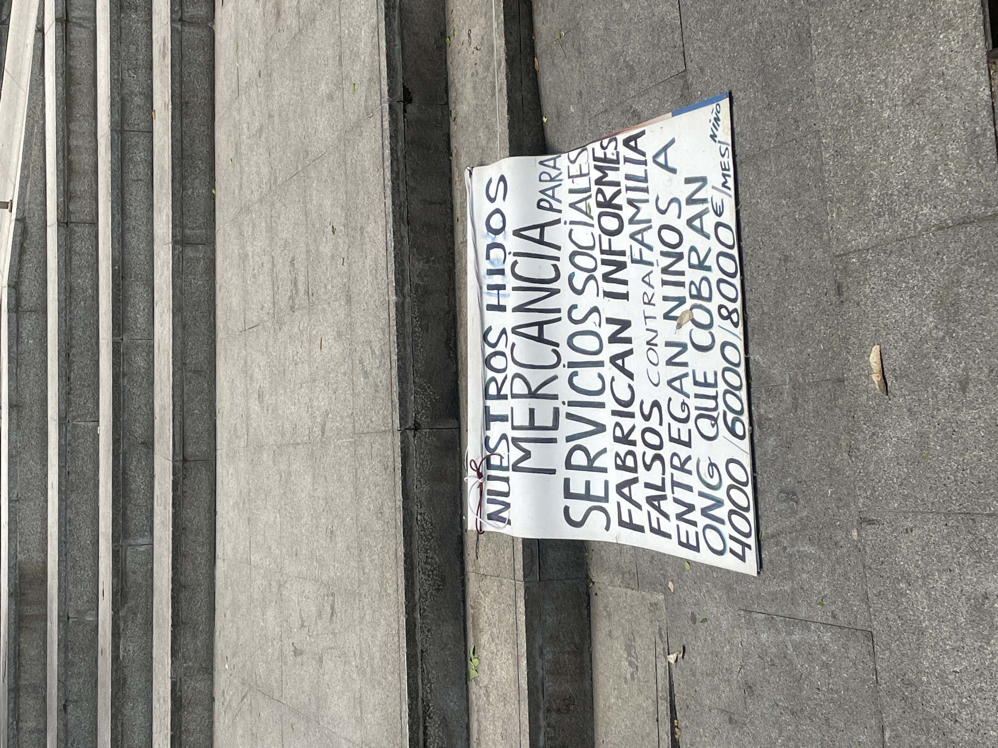 A sign is lying on the ground next to a set of steps. The sign is written in Spanish and the text translates to "Our children merchandise for social services fabricate false reports against family deliver children NGO that charges 4000/6000/8000/month". The sign appears to have been placed on the ground near the steps. The background suggests a city environment. The photograph depicts a controversial message about NGOs.