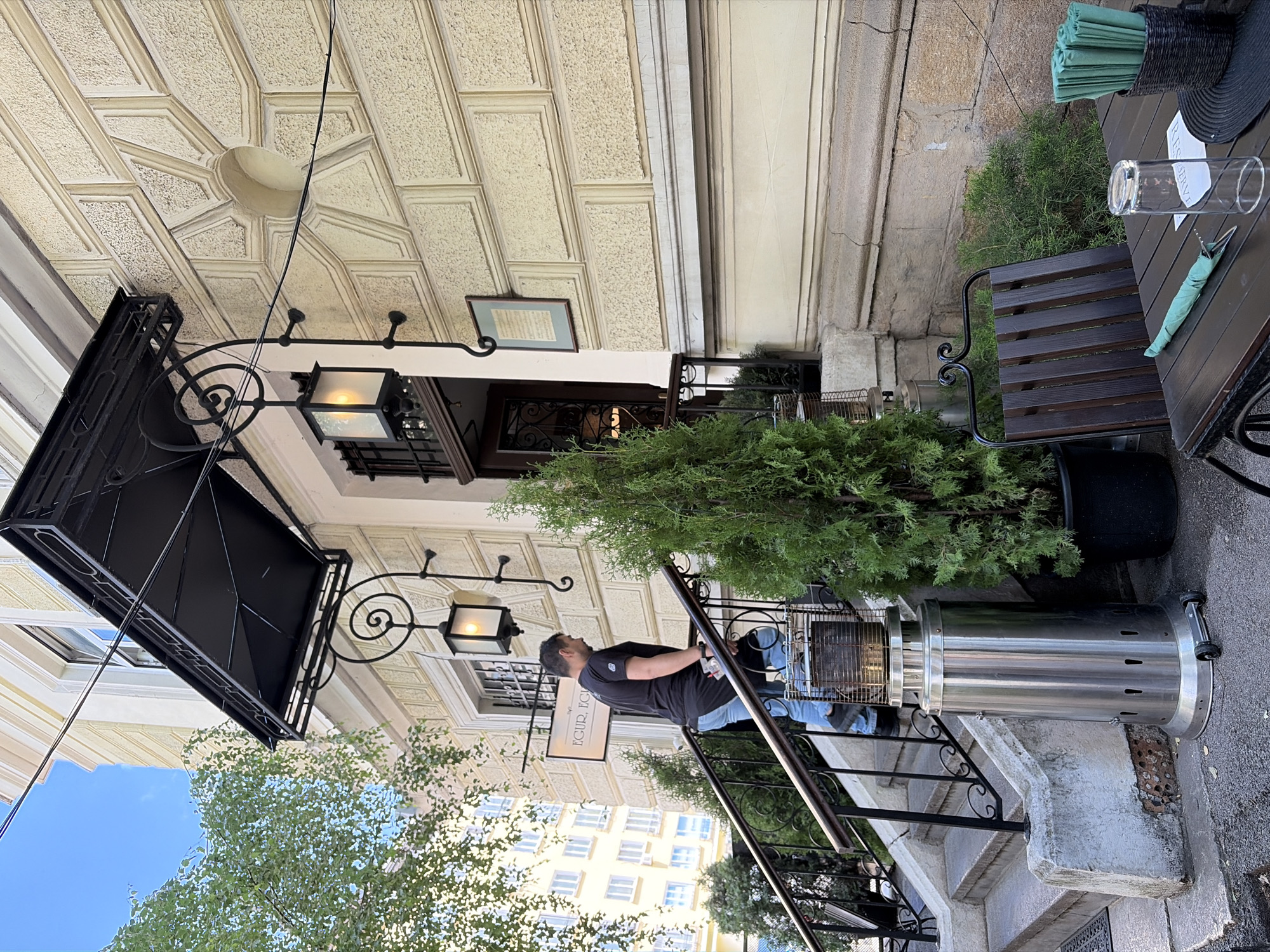 The image captures an outdoor seating area of a restaurant, likely in Sofia, Bulgaria, during the daytime. A man in a black t-shirt and blue jeans stands on a set of stairs leading up to the entrance, looking towards the building. He appears relaxed. To the right, a table is set for dining with a clear glass, a folded green napkin with cutlery, and a stack of green napkins in a woven holder. A "RESERVED" sign is placed on the table.
The architecture of the building features an ornate, textured ceiling with geometric patterns, and a black metal awning above the entrance. An old-fashioned streetlamp with a frosted glass shade hangs from an ornate bracket. Green bushes and trees are visible, providing a natural element to the urban setting. In the background, a building with many windows suggests a city environment. A tall, cylindrical stainless steel patio heater stands to the right of the stairs, indicating a potentially cooler time of day or season, or simply for ambiance. The overall impression is a pleasant, perhaps slightly sophisticated, outdoor dining space. The visible text on a sign reads "EGUR. EGURI".