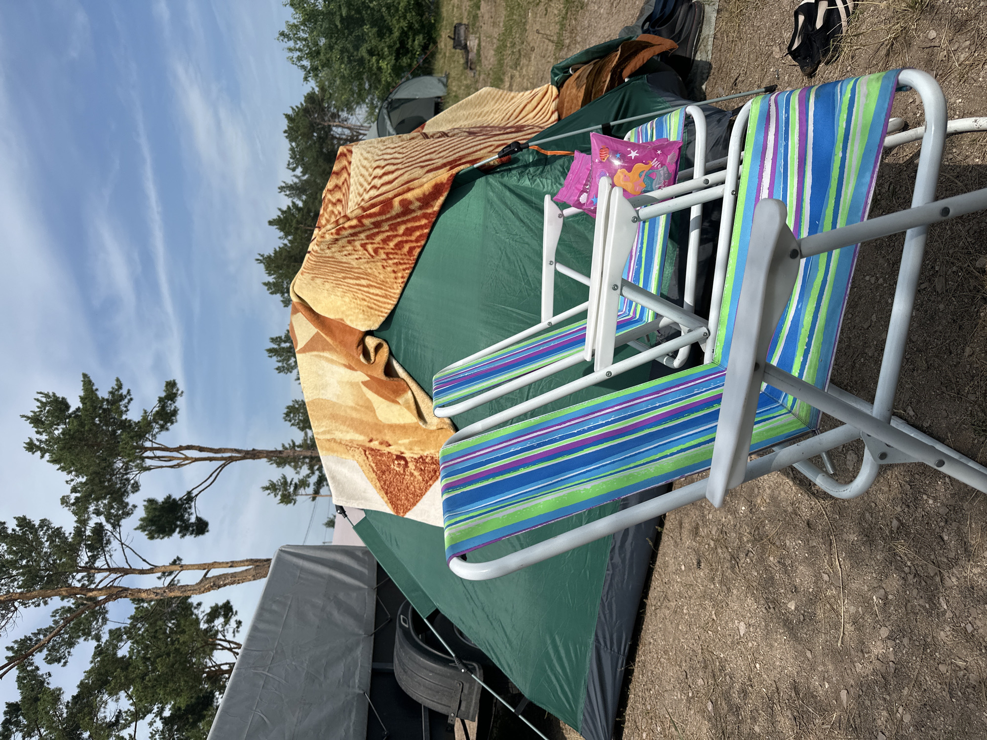 In the foreground of the image, two colorful striped folding chairs sit on a dirt and gravel campsite. Behind the chairs is a green tent with a patterned blanket draped over its top. To the left of the tent, a portion of a gray tarp-covered trailer is visible. Trees and a blue sky with scattered clouds form the background. The scene depicts an outdoor campsite, likely in the city of Kolodezny, Russia.