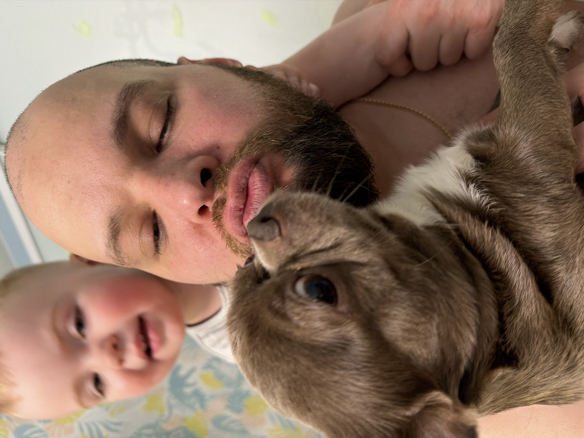 A bald man with a beard lies sideways, eyes closed, holding a brown and white dog close to his face. He is wearing a gold chain. A small child is visible to the left. The background appears to be a patterned sheet. The location is unknown and there is no evidence to suggest the scene is in Votkinsk, Russia.