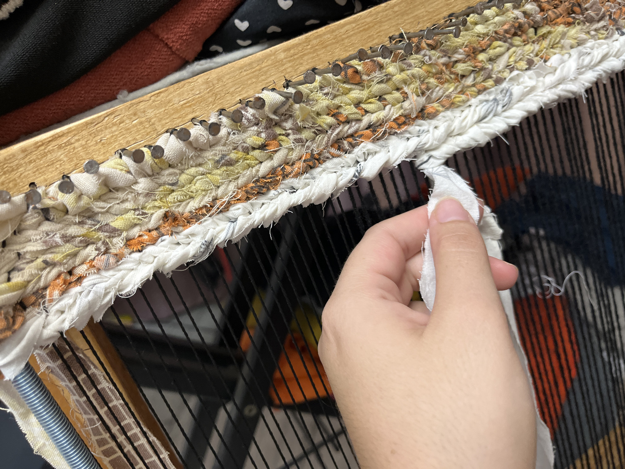 An overhead, close-up shot shows a person's right hand holding a strip of white fabric to a loom. The loom frame is made of light-colored wood, and the warp threads are black. The weft material is made of braided fabric strips in various colors, including white, cream, yellow, orange, and brown, and is attached to the loom by small nails. Behind the loom, a dark, patterned fabric is visible, possibly the background or other materials for crafting. The scene is indoors, and the lighting is natural. The location is not discernible from the image.