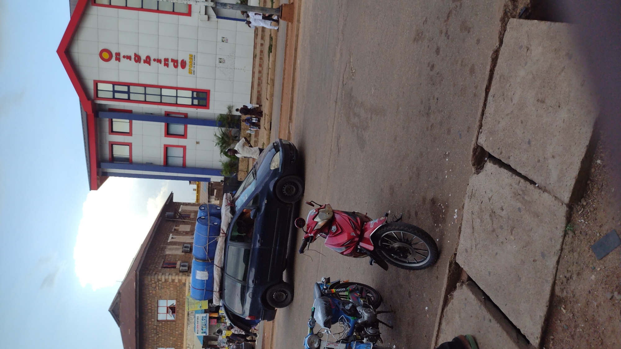 In Jos, Nigeria, a dark blue minivan is parked on a paved road, its rear door open. Blue cylindrical containers are secured to the roof of the minivan. Two motorcycles, one blue and one red, are parked to the left of the minivan on the road. A modern building with white facade and red trim is visible in the background to the right, with the vertical text "Omoigbinde" on its side. To the left of the modern building, a brown brick building with a sloping roof is present. Several people are walking and standing on the sidewalk in front of the buildings. The sky is overcast, with large white clouds.