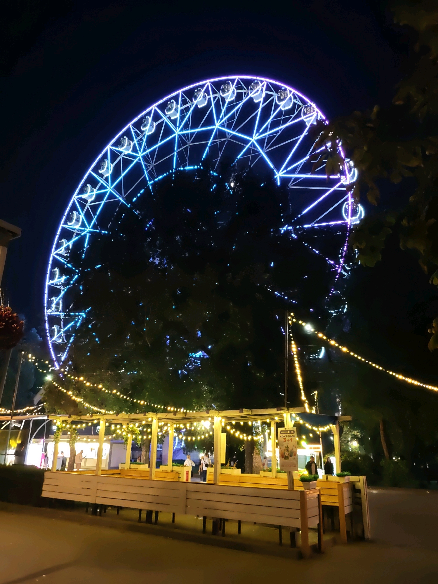 The image depicts a Ferris wheel illuminated with blue and purple lights at night. In the foreground, a wooden structure with benches and a roof adorned with string lights is visible. Several people are standing around this structure, some appearing to be patrons or staff. The scene suggests a fairground or amusement park setting, possibly at dusk or night. Based on a sign in Russian which reads "КОКТЕЙЛИ 3+1" (Cocktails 3+1), the location is identified as Rostov-on-Don, Russia, specifically at the Rostov-on-Don amusement park, located on the Don embankment.
