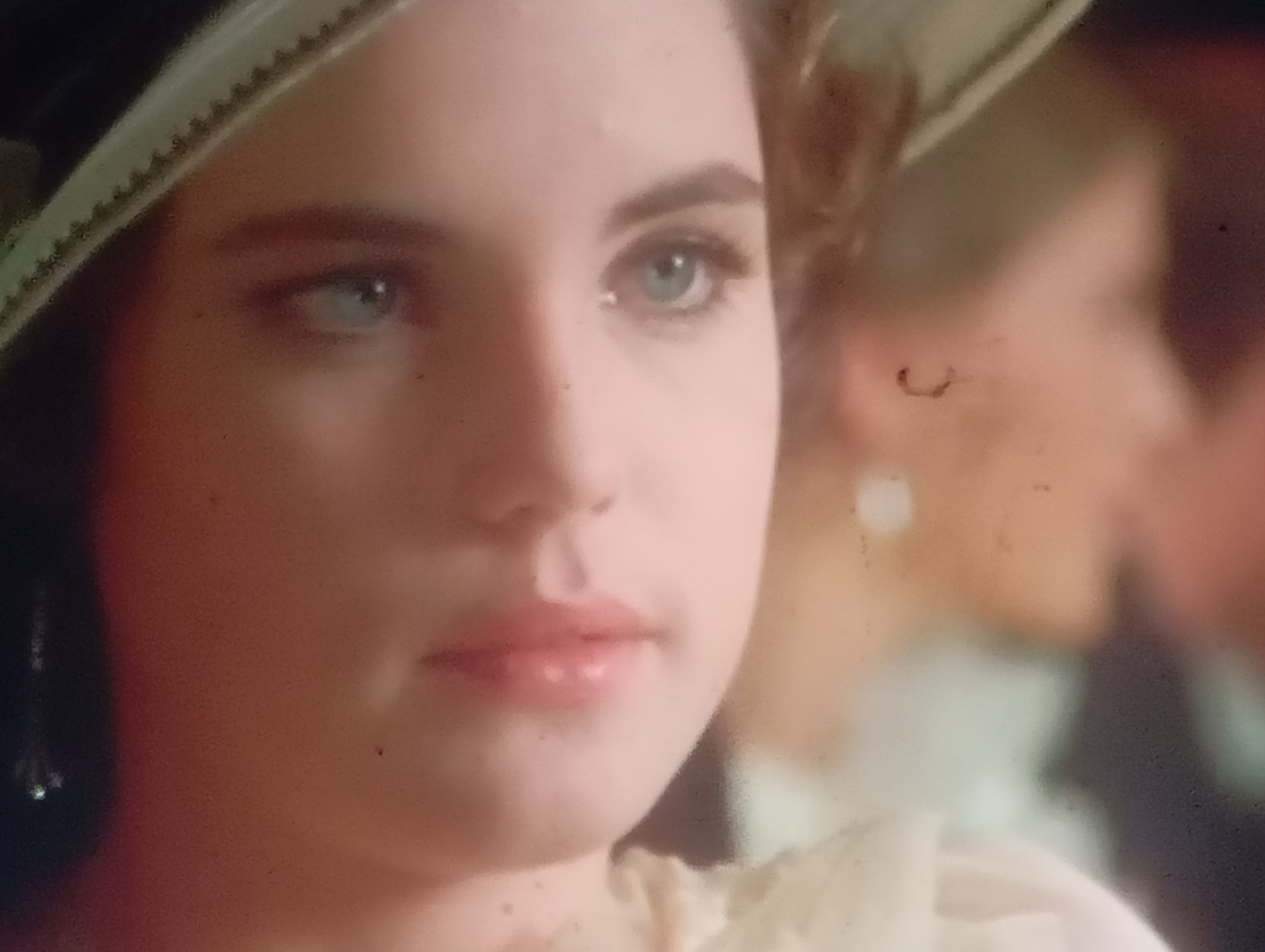The image is a close-up, out-of-focus shot of a young Caucasian woman's face, with her head tilted slightly to the right. She is wearing a hat with a wide brim that has a decorative, ridged edge. Her left eye, which is lighter than her right, is clearly visible and appears to be light blue or green. She has dark eyebrows and appears to be wearing some makeup, including lipstick. The background is blurred, showing hints of other people or objects, suggesting an indoor event or gathering. The lighting is soft, with a warm tone. There is no indication of the city of Sofiivska Borshchahivka, Ukraine, or any specific location within it.