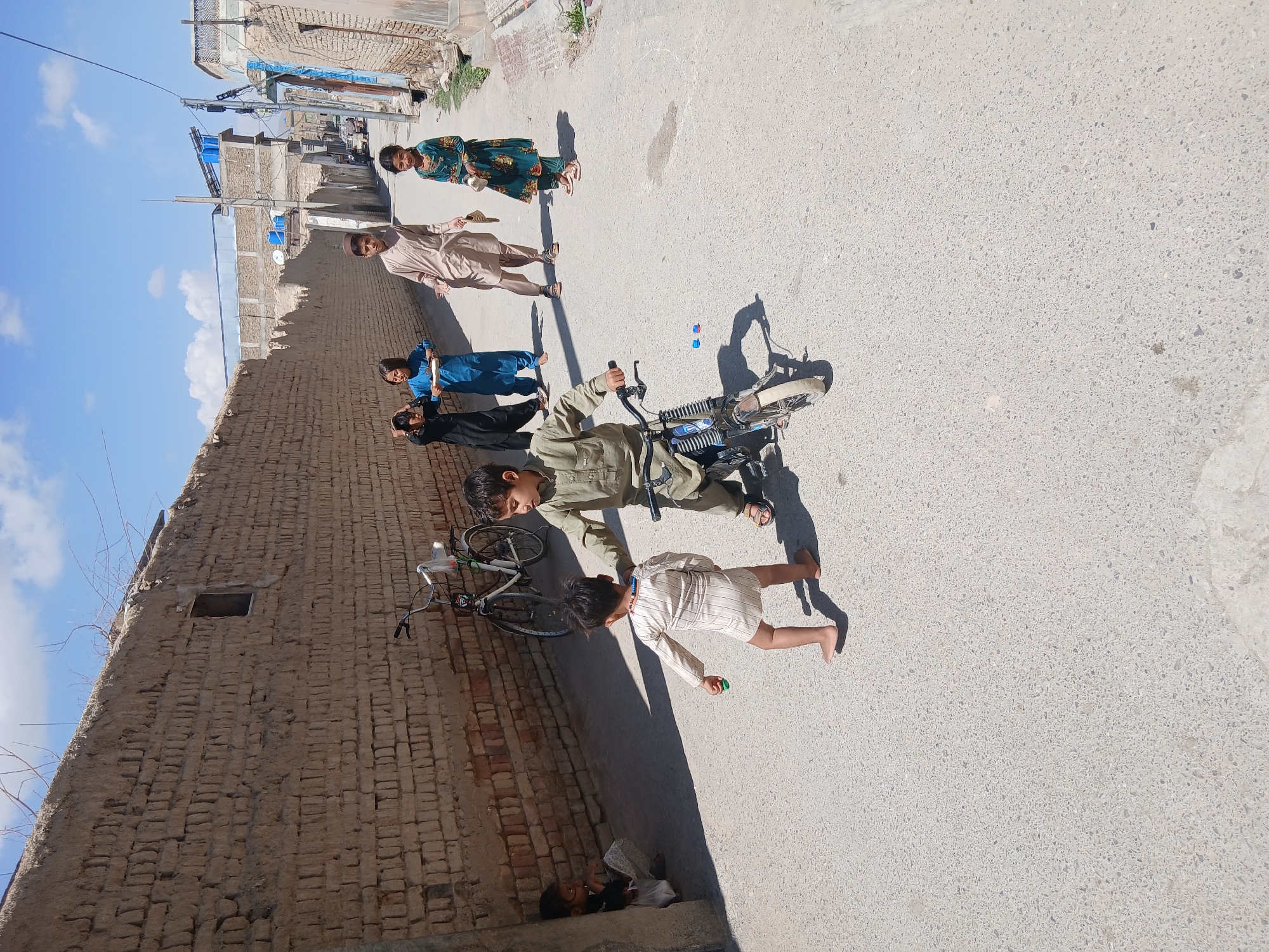 The image depicts a street scene in Quetta City Tehsil, Pakistan. A group of children is present on the road. One child is riding a small motorcycle, while another runs alongside it. Further down the street, other children are walking. The environment consists of a brick wall on one side and buildings lining the other side of the street. The weather appears to be sunny.
