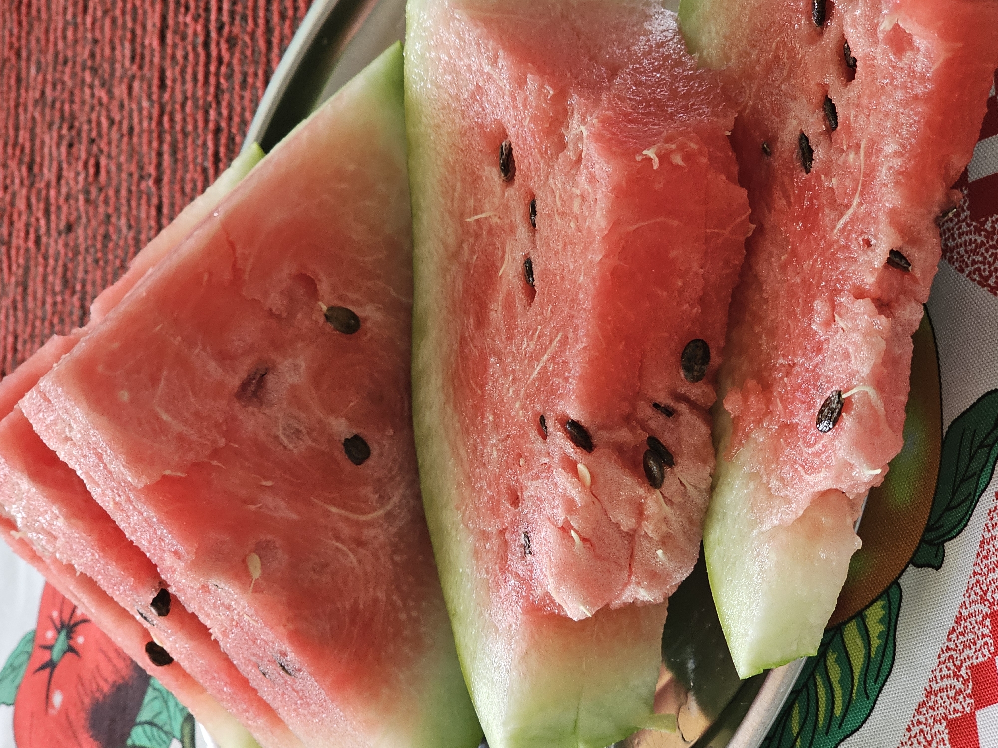 The image displays several triangular slices of ripe watermelon, stacked on a metal tray. The watermelon flesh is a vibrant pink, speckled with black seeds. The rind is a pale green, fading to white near the flesh. The slices appear juicy and fresh. The tray is placed on a patterned tablecloth. In the background, a textured red surface is visible. The scene is indoors. The location of Özgön, Kyrgyzstan is not depicted or suggested in the image.