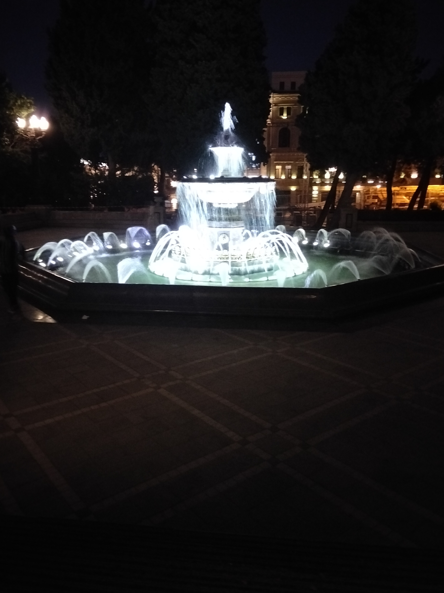 The image captures a lit-up fountain in a city square at night. The fountain is the central subject, with multiple tiers of water spraying upwards and outwards, illuminated by bright white lights. Around the fountain, there are sprays of water creating arcs and cascades. In the background, trees with dark foliage are visible, along with the facade of a multi-story building that appears to be illuminated with yellow lights. Streetlights with warm yellow glows are also present to the left, and a street lamp is visible between the trees. The ground in the foreground is paved with square tiles, with diagonal lines creating a patterned effect under the lighting. A person is partially visible on the far left of the image, appearing as a dark silhouette. The overall scene depicts a public urban space at night, likely a park or plaza, in Sabail Raion, Azerbaijan.