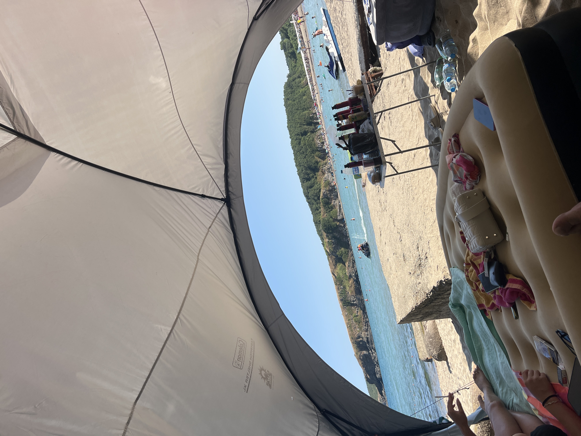 The image depicts a beach scene in Sinemorets, Bulgaria, viewed from inside a white canopy tent. In the foreground, an inflatable mattress is set up on the sand with various items, including towels, a purse, a phone, and a pack of cigarettes. To the right of the mattress are several plastic water bottles.
Further back, a portable folding table is laden with bottles, cups, and a black trash bag. Behind the table, the sandy beach extends to the clear blue sea, where several people are visible. There are jet skis and a boat on the water. The coastline is lined with lush green trees and hills. The sky is a bright, clear blue, indicating a sunny day. The overall impression is one of a relaxed beach outing.
