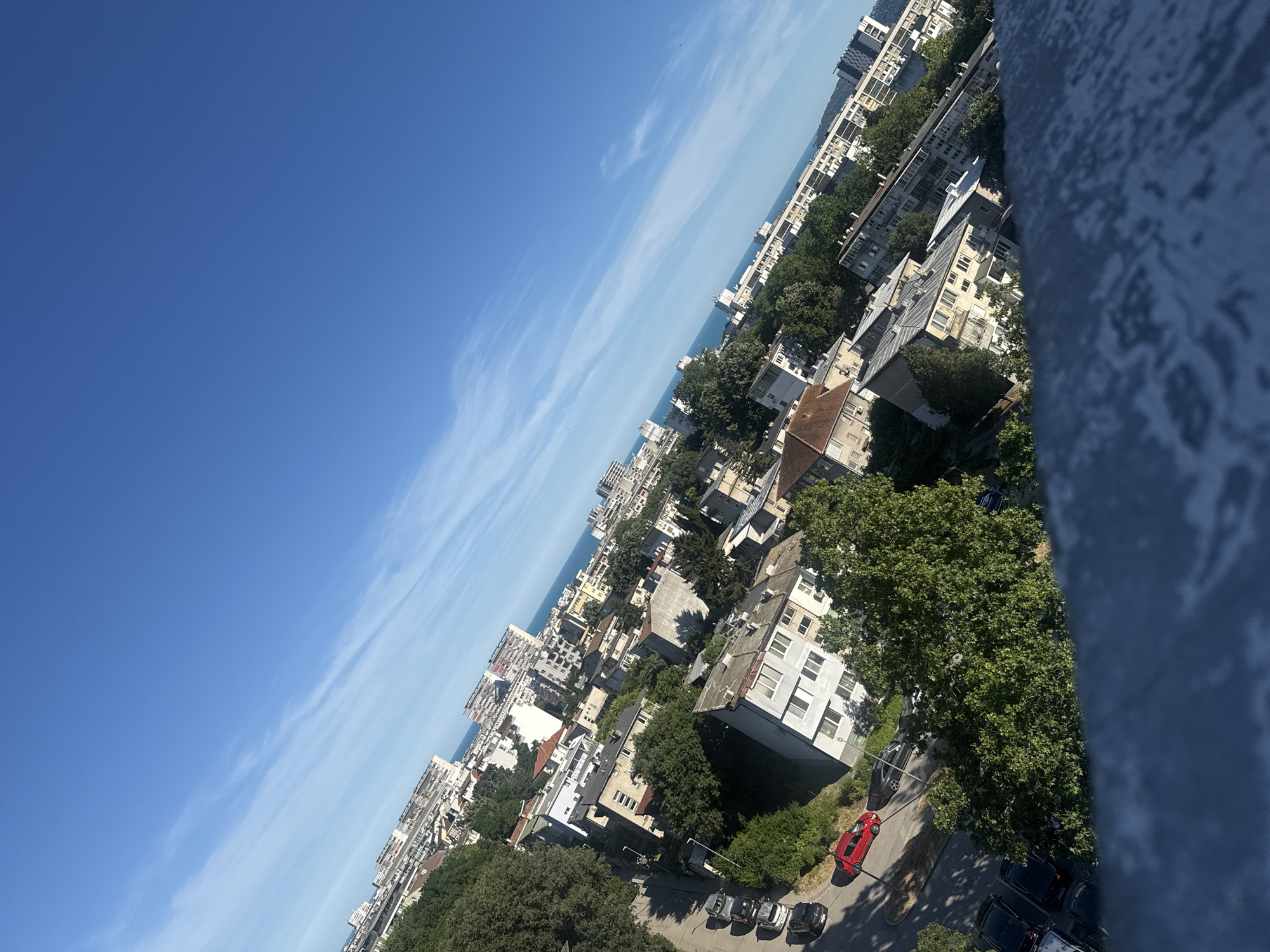The image depicts an aerial view of a city, likely Varna, Bulgaria, on a clear day with a vibrant blue sky. The scene is dominated by a dense urban landscape characterized by numerous residential buildings, varying in architectural style and height, interspersed with lush green trees. The buildings appear to be mostly mid-rise, with flat roofs and visible balconies.
In the foreground, a textured, possibly concrete surface, tilted at an angle, occupies the bottom portion of the frame. Further into the scene, a street is visible with several parked cars, including a bright red car and several darker colored vehicles. The street curves and is lined with trees, casting shadows on the pavement.
Beyond the immediate urban area, a glimpse of the sea or a large body of water can be seen in the distance, suggesting a coastal location. A hazy horizon is formed by more buildings stretching along the coastline. The overall impression is one of a typical residential neighborhood within a coastal city, captured from an elevated perspective. There are no people clearly visible in the image.
