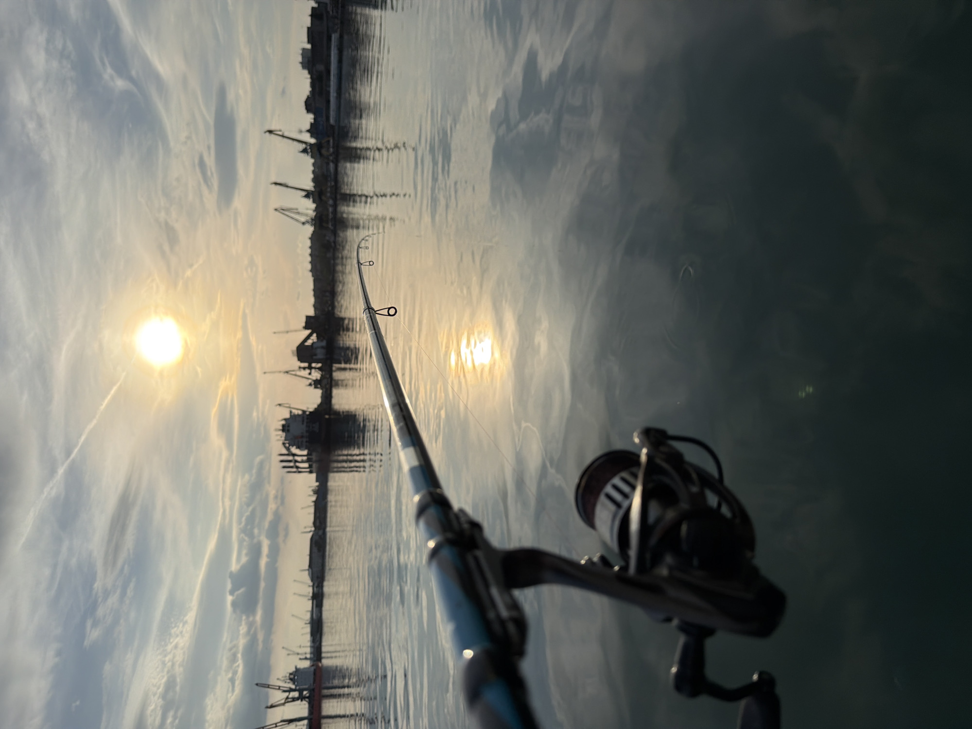 The image shows a fishing rod and reel in the foreground with the cityscape of Burgas, Bulgaria, in the background. The sun is setting, casting a reflection on the water. Cranes and buildings line the coast. The water appears calm. The fishing line extends into the water. No people are visible.