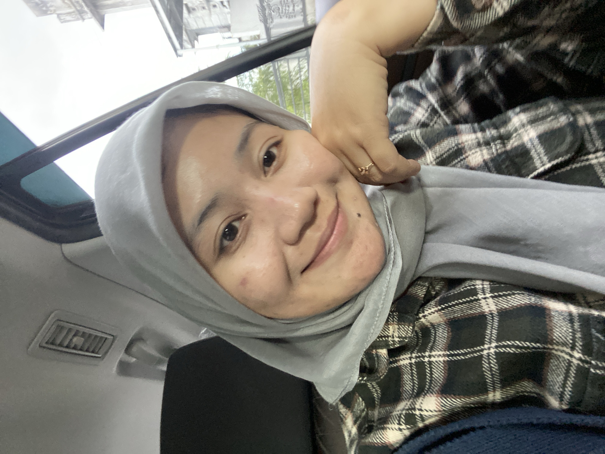 A young Indonesian woman wearing a grey hijab and a plaid shirt poses for a selfie inside a car. The woman is leaning her head on her hand, with a ring on her finger. Behind her, the interior of the car is visible, including a ventilation grate and part of a seat. Outside the car window, buildings and trees can be seen, suggesting an urban environment in Banjarbaru, Indonesia. The lighting is natural, indicating it is daytime.