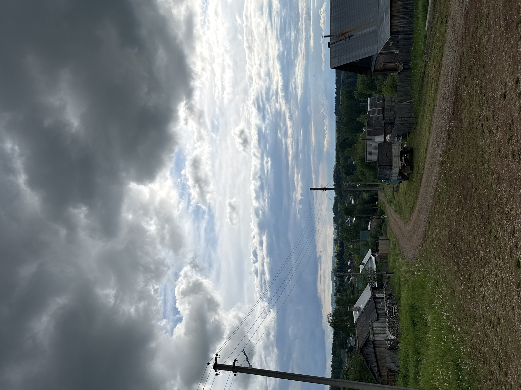 The image presents a rural village scene in Бородулино, Russia, under a dramatically changing sky. The upper left portion of the frame is dominated by heavy, dark grey storm clouds, while the sky transitions towards the upper right to reveal brighter, streaky white clouds and patches of clear blue sky.
A dirt road or track runs from the foreground along the right side, curving slightly into the distance. Lush green grass, dotted with small white flowers, borders the road on the left. Several rustic wooden buildings are scattered throughout the village. A large wooden house with a dark metal roof stands prominently on the far right. Further along the road, other traditional wooden structures are visible, with a dark-colored truck parked next to one in the mid-ground.
A dense line of green forest forms the horizon in the background. Wooden utility poles carrying power lines traverse the mid-ground, adding an element of infrastructure to the natural landscape. The scene appears quiet and devoid of people or specific activities. The lighting suggests it is daytime, possibly late afternoon, given the interplay of bright and dark clouds. No visible text is present in the image.