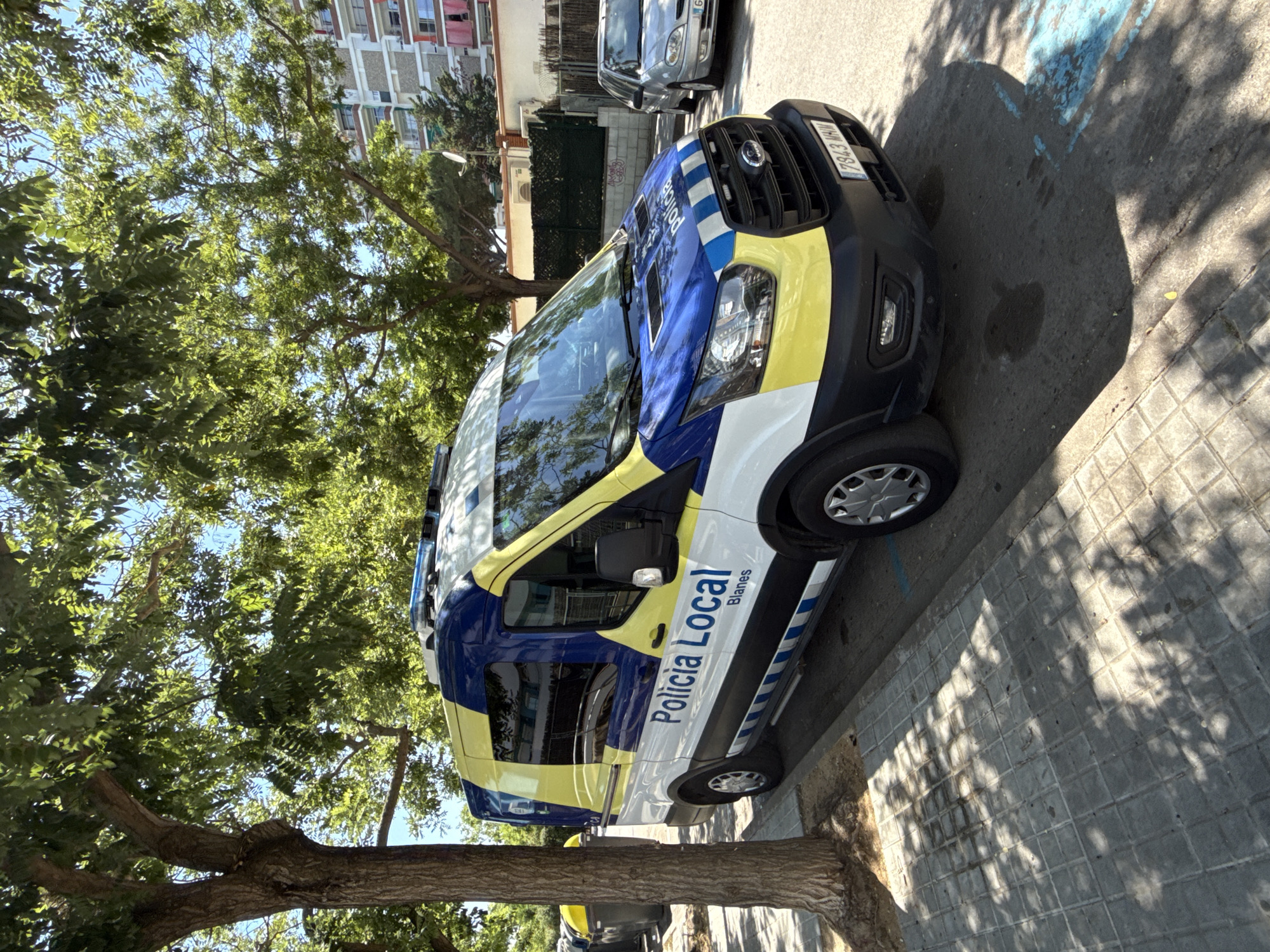 A police van is parked on a tree-lined street in Blanes, Spain. The van is white, blue, and yellow, with "Policia Local Blanes" written on the side. A silver car is parked behind the police van. The street is paved with gray tiles, and there are shadows from trees on the pavement. In the background, there is a multi-story building and a fence. A trash can is visible on the left side of the image. The sky is blue with some clouds.