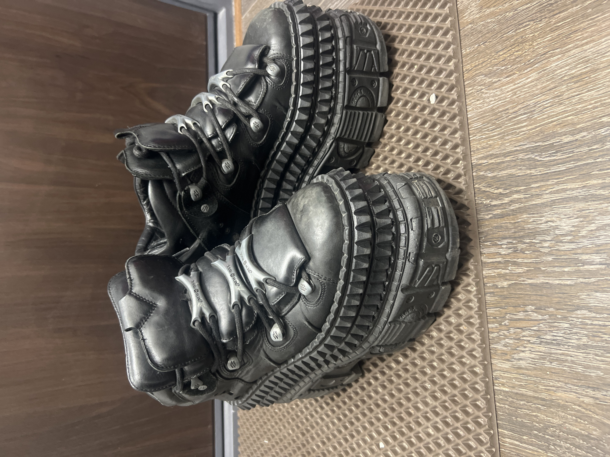 A pair of black, heavily-soled platform boots is centered in an indoor setting. The specific geographical location, including the city of Unknown, Russia, cannot be determined from the visual evidence. The boots feature high-top construction with black laces, multiple silver-colored eyelets, and metallic lace bars embossed with "NEW ROCK" branding. Their prominent platforms display multi-layered designs, intricate molded patterns, and a perimeter of aggressive, conical studs or spikes. The leather-like material of the boots appears worn, showing scuffing and dust, particularly on the toe areas and platforms. The footwear rests on a light brown, waffle-textured rubber mat that abuts a wooden floor with a distinct linear grain. A darker brown wooden wall or paneling is visible in the background. No individuals are present, and no actions are depicted.