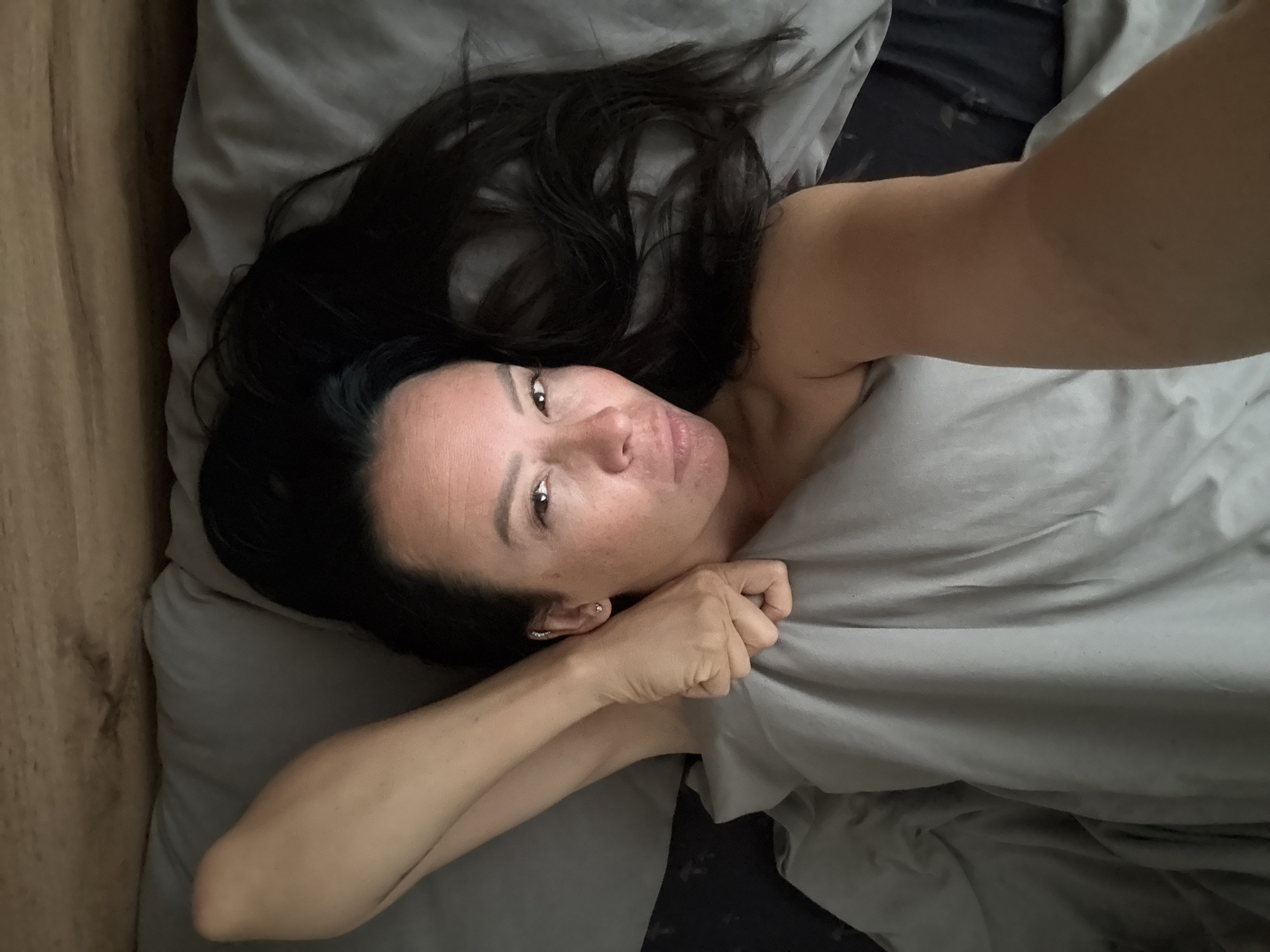 An overhead shot captures a woman lying in a bed with gray sheets. Her head is tilted, and her dark hair is spread out. One arm is raised, bent at the elbow, and her hand is near her head. Her other arm is extended to the left, partially covered by the sheet. The woman is looking towards the camera with her eyes open. A wooden headboard is visible to the right of the bed.