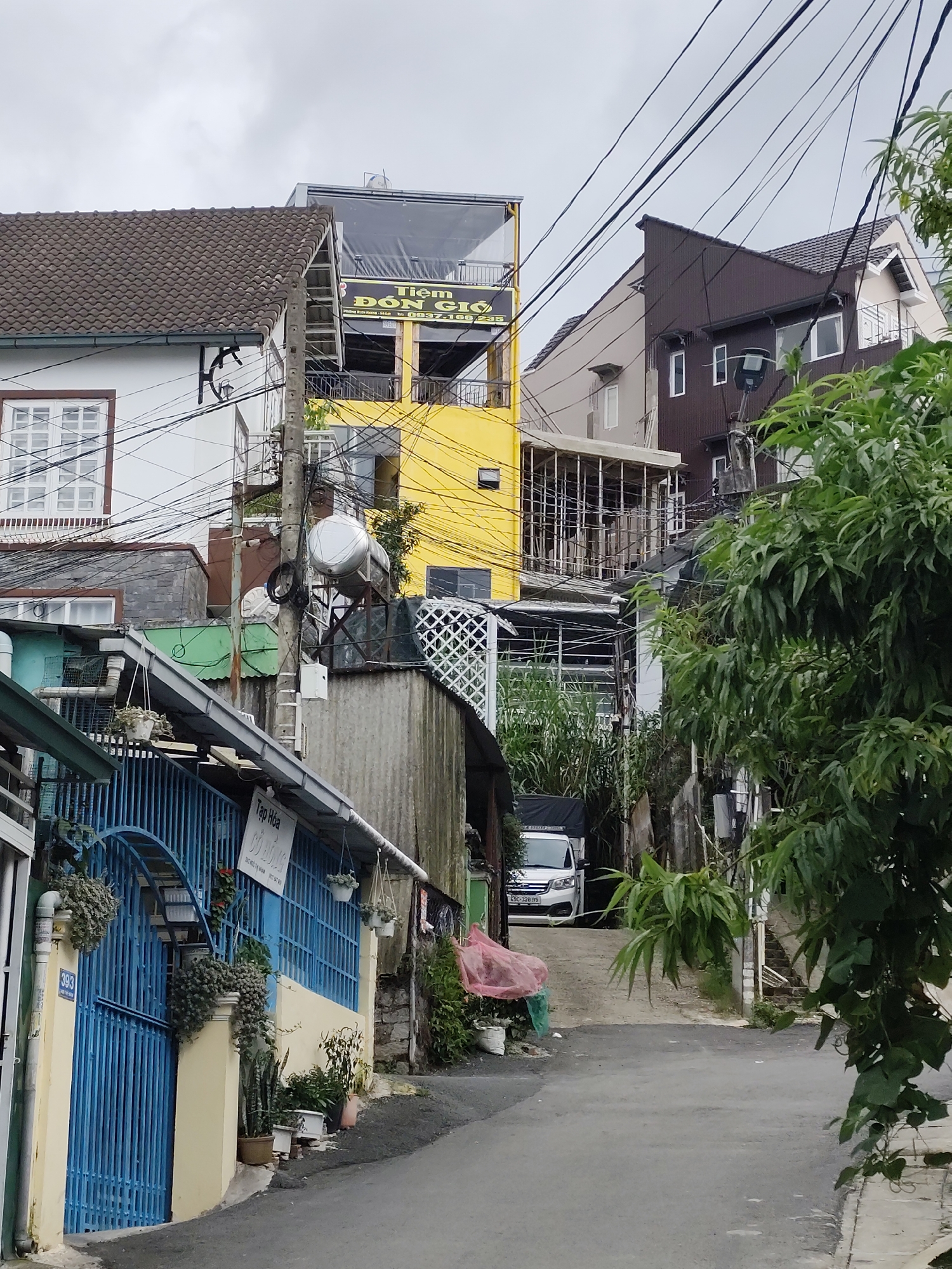 The image depicts a narrow street in Da Lat, Vietnam, lined with multi-story buildings. The architecture is varied, with some structures featuring tiled roofs and others with more modern facades. Power lines are prominently visible, crisscrossing overhead and contributing to a sense of urban density.
The building on the left is white with a dark-tiled roof and a large window with white panes. Adjacent to it is a bright yellow building with multiple balconies and signage that appears to be in Vietnamese. To the right of the yellow building, a darker, multi-story structure with a pitched roof is visible, also with several windows.
In the foreground, a blue metal gate with decorative scrollwork leads into a property. This gate is adjacent to a cream-colored wall. Plants and hanging baskets add a touch of greenery to the scene. Further down the street, a white van is parked. The road surface is asphalt. The overall impression is of a residential or mixed-use area in a Vietnamese city, characterized by a mix of building styles and the ubiquitous presence of electrical infrastructure. The sky is overcast, suggesting a cloudy day.
