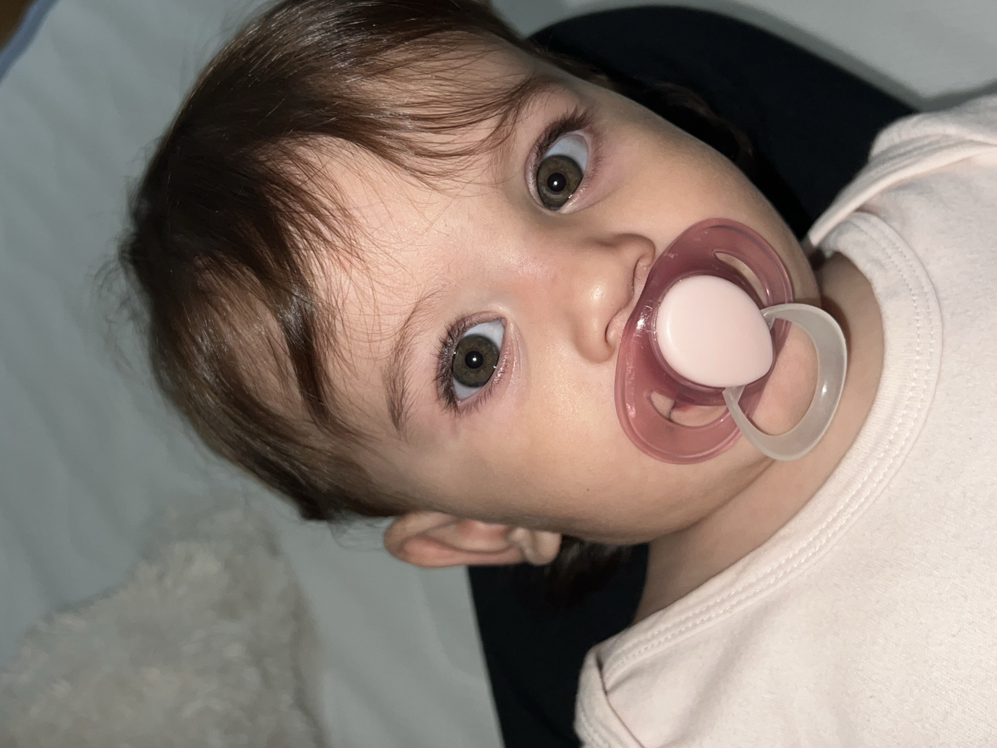 A young child with brown hair and green eyes is looking directly at the camera while holding a pink pacifier in their mouth. The child is wearing a light pink shirt. The background includes a white blanket and a dark-colored garment. The location is not discernible from the image.