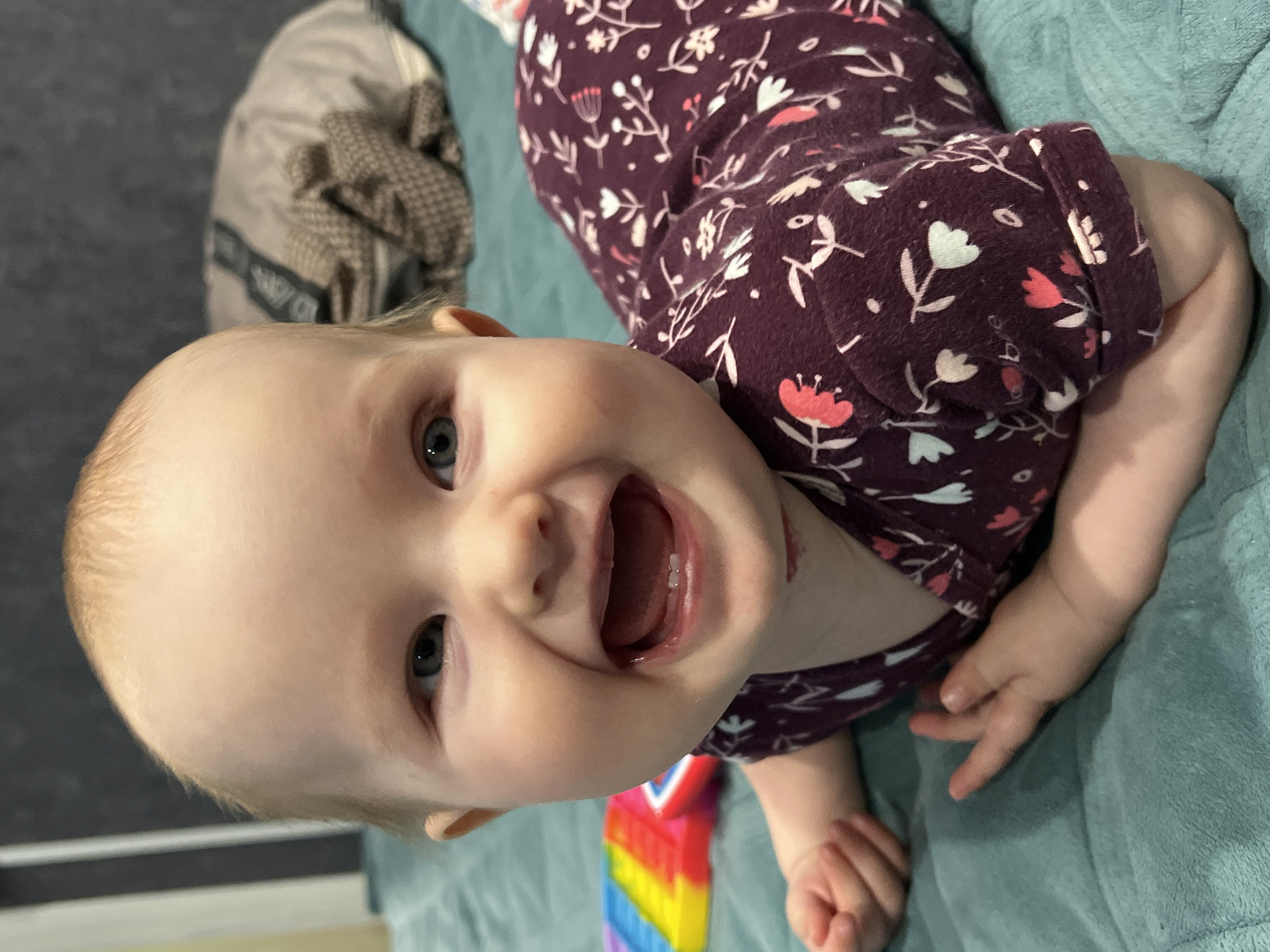 A baby is lying on a teal blanket, wearing a long-sleeved, burgundy patterned top. The baby is smiling, and two bottom teeth are visible. The baby is on a light-colored surface with a rainbow-colored toy partially visible. Behind the baby is a tan bag or object. The location is unknown and unrelated to Chaikovsky, Russia.