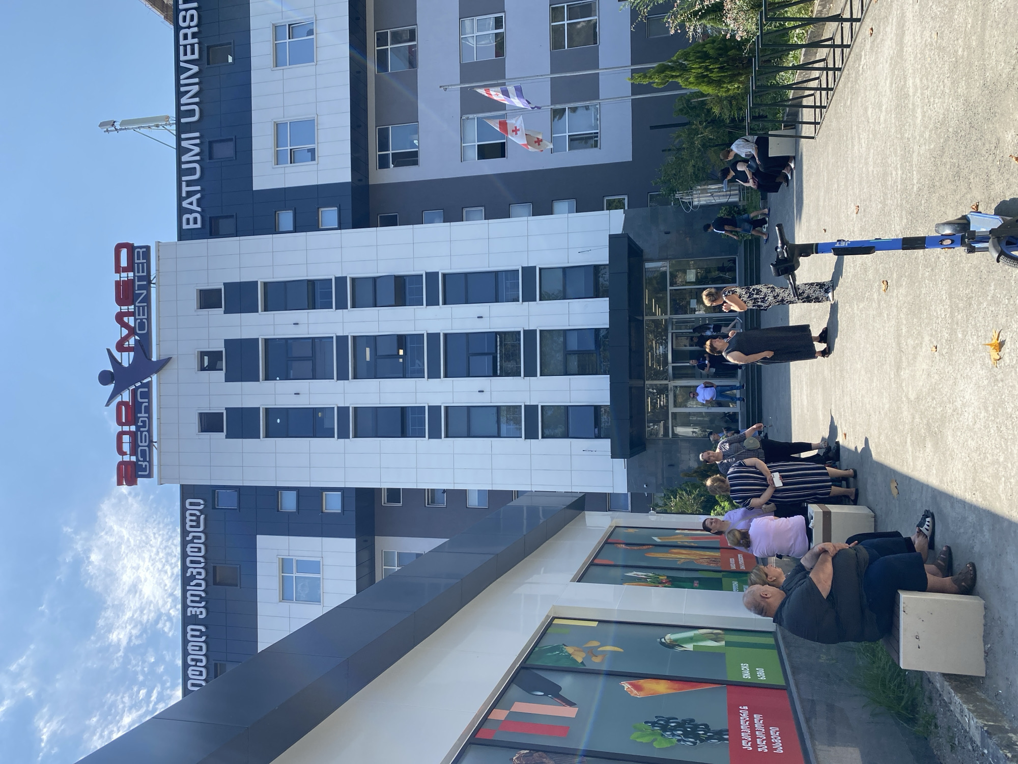 The image shows the Batumi University building in Batumi, Georgia, on a sunny day. The building has a modern facade with dark blue and white panels. The words "BATUMI UNIVERSITY" are visible in large white letters on the building. A smaller sign with red lettering that says "MED CENTER" is also visible at the top of the building, along with a stylized red logo of a person.
In front of the building, several people are standing and sitting. Some are gathered near the entrance, while others are seated on benches or waiting in line. A blue electric scooter is parked on the pavement in the foreground. The environment appears to be an urban setting with trees and foliage around the building. Two Georgian flags are flying from poles in front of the building. A large mural or advertisement is displayed on the side of an adjacent building to the left.