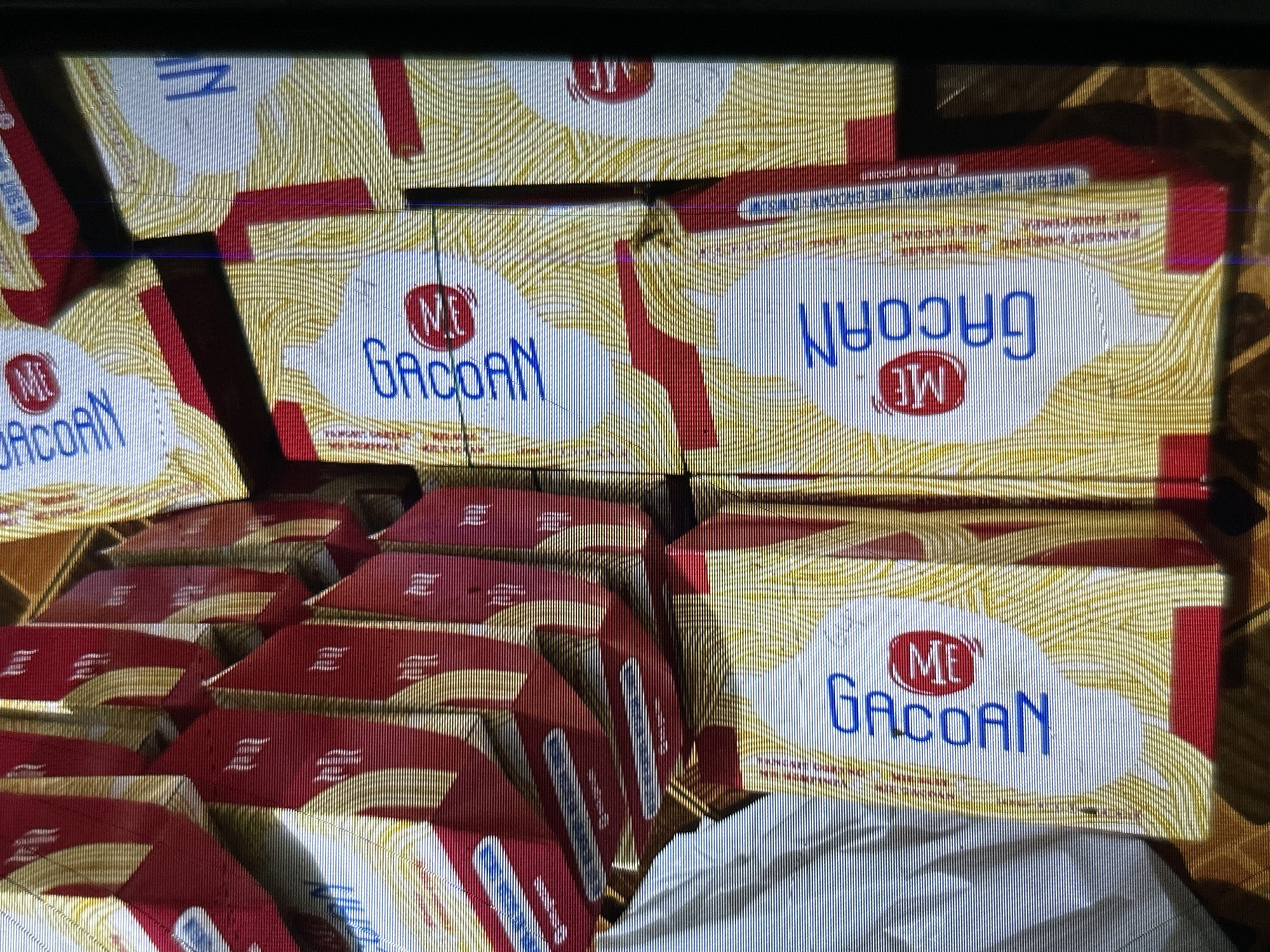 The image shows a stack of spaghetti boxes, likely in a store or warehouse, in Depok, Indonesia. The boxes are predominantly red and white with yellow detailing depicting spaghetti strands. The brand name "GACOAN" is visible in blue lettering on some of the boxes, along with a red circular logo containing the letters "ME". The lighting is dim, and the image appears to be taken from a slightly elevated angle. The boxes are arranged in a somewhat haphazard manner, suggesting they are in storage or awaiting display.