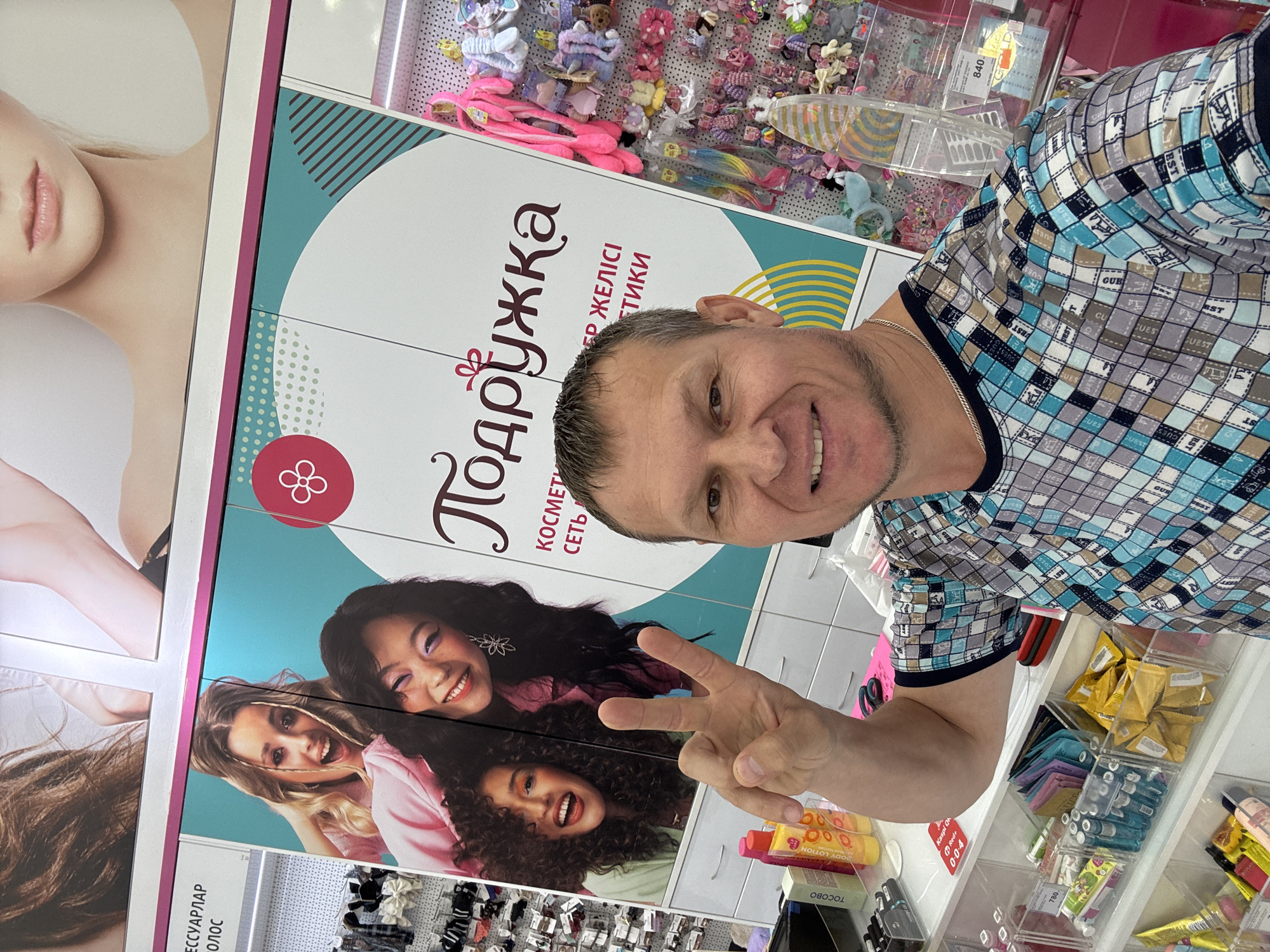 A man with short brown hair and stubble is posing for a selfie in what appears to be a retail store. He is wearing a checkered shirt with blue, gray, and white patterns. The man is smiling and holding up two fingers in a peace sign. Behind him, there is a large poster with images of three women, one with blonde hair and two with dark hair, smiling. The poster has text in Russian, including "Подружка" which translates to "Girlfriend". To the right of the poster, there are shelves stocked with various hair accessories and cosmetics. The store appears to be well-lit and organized. Based on the context of the Russian text and the style of the store, it is likely located in a Russian-speaking region. While the specific location in Oskemen, Kazakhstan cannot be definitively confirmed from the image alone, the presence of Russian text suggests a connection to a region where Russian is commonly used.