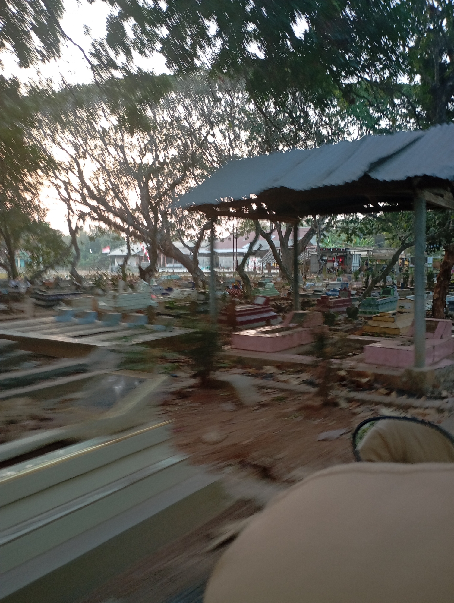 The image depicts a cemetery in Kecamatan Haurgeulis, Indonesia, with numerous gravesites visible. The graves are varied in style and color, with some featuring raised concrete structures and others appearing to be simple mounds. A corrugated metal roofed shelter stands in the middle ground, providing shade over a few of the graves. Mature trees with dense foliage surround the cemetery, and the sky is visible through their branches, suggesting it is either early morning or late afternoon due to the soft lighting. In the background, several buildings with tiled roofs can be seen, and a Indonesian flag is flying. The overall impression is one of a quiet, well-maintained resting place.