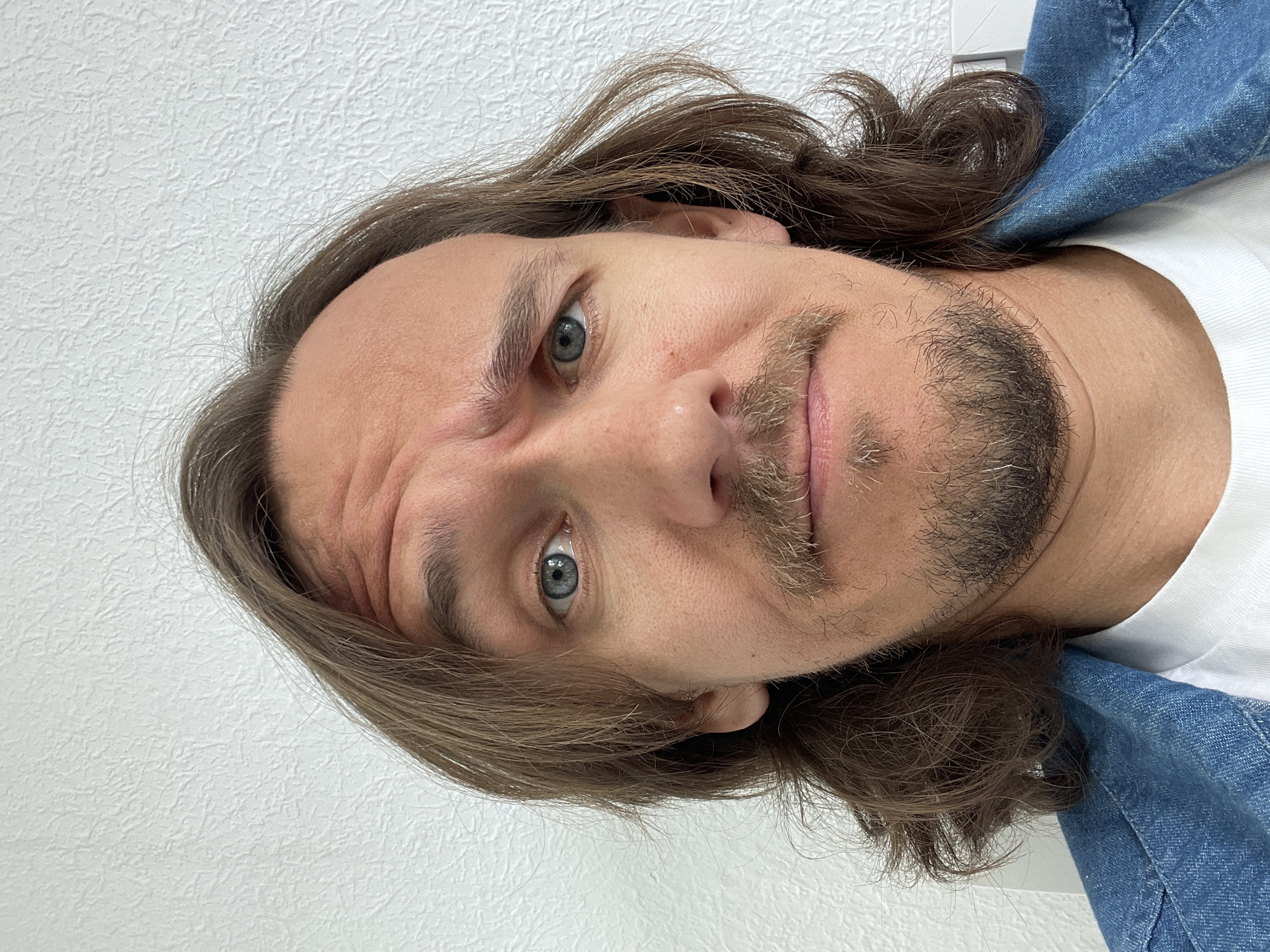 The image displays a close-up portrait of a man, originally captured with a head tilt and rotated ninety degrees to the left. He has long, wavy brown hair that cascades around his face and neck. His light blue eyes are wide and focused directly on the viewer, conveying a curious and slightly playful expression. A light brown mustache and goatee, interspersed with some grey hairs, frame his mouth, which holds a subtle, almost smirk-like curve.
He is wearing a white t-shirt underneath a blue denim jacket, visible on his right shoulder and chest area. The background is a plain white wall with a distinct textured pattern, suggesting an indoor setting. There are no other objects, visible text, or specific location cues. The lighting appears even, typical of an interior space, and the time of day is not discernible. The man's direct and engaging gaze indicates he is looking at the camera, likely taking a selfie.