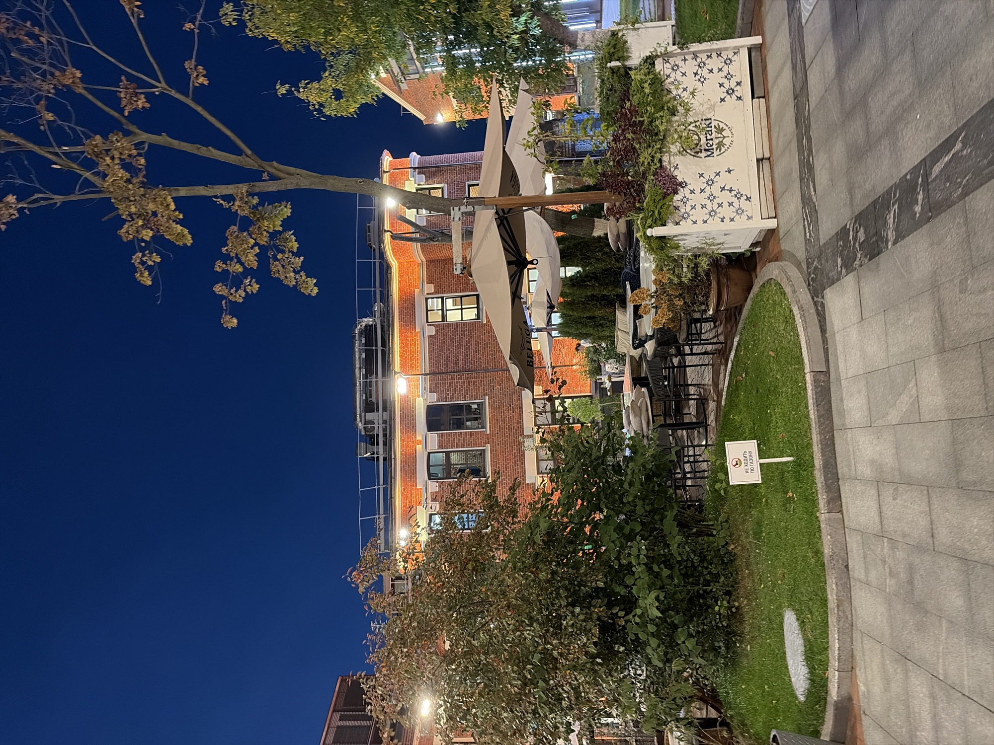 This image depicts the exterior of a restaurant in Moscow, Russia, at dusk. The building, constructed of red brick, features multiple windows and is illuminated by warm light emanating from within and from decorative string lights along the roofline. In the foreground, an outdoor seating area is visible with tables and chairs, shaded by large, cream-colored umbrellas. A planter box with decorative blue and white patterns, bearing the text "MERAKI GREEK TAVERN," is positioned on the right. Adjacent to the seating area is a patch of green grass with a small "No Walking on the Grass" sign written in Cyrillic. Several trees are present, one with browning autumn leaves and another with lush green foliage. The sky is a deep blue, indicating the late hour. No people are clearly identifiable in the scene.