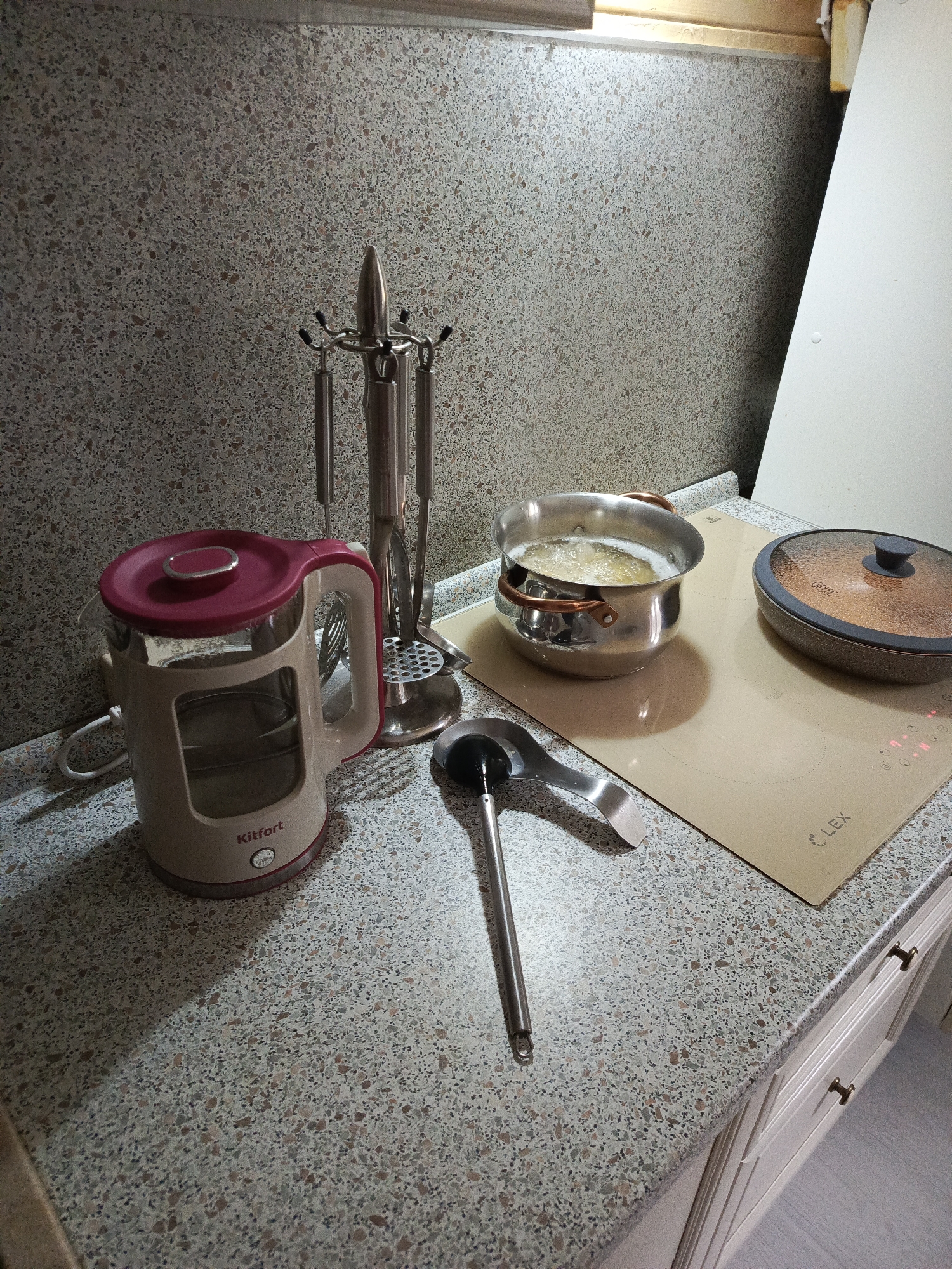 The image shows a kitchen countertop with various items arranged on it. A red and beige electric kettle stands to the left, next to a stainless steel utensil holder with hanging utensils. A pot filled with boiling liquid sits on a beige-colored induction cooktop, along with a covered pan. A ladle lies on the countertop in front of the pot. The countertop has a speckled gray and beige pattern. The background includes a wall with a similar speckled pattern and a white cabinet. The city of Северный, Russia cannot be determined from the image.