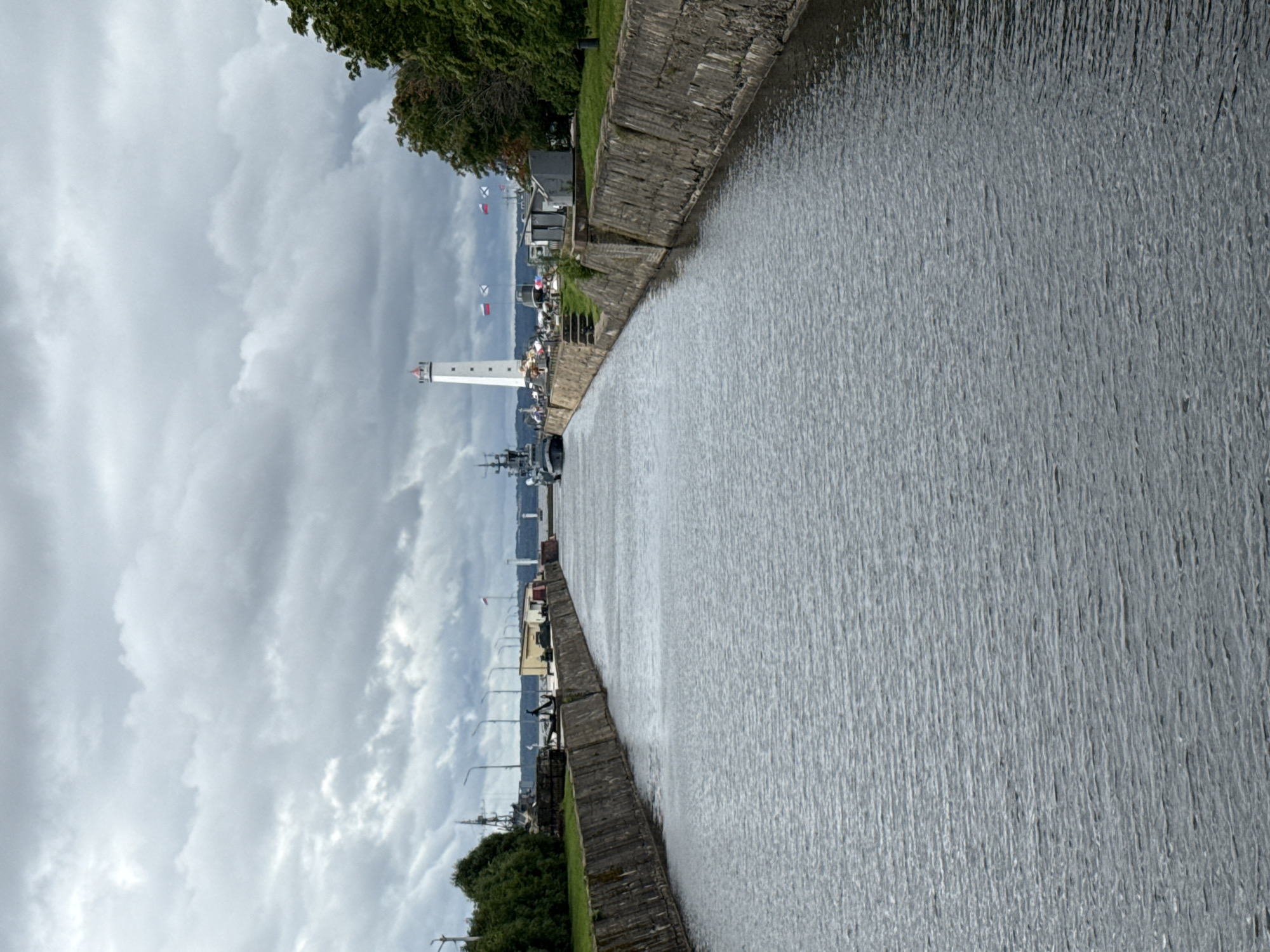 This is a wide-angle, vertical shot of a canal in Kronstadt, Russia. The canal is lined with weathered concrete walls on either side. The water in the canal is dark and choppy, reflecting the cloudy sky. In the background, a white lighthouse with a red top stands tall. Several ships are docked along the canal, including a large gray vessel to the left. Flags, some appearing to be Russian, are flying from poles along the embankment. Trees and grassy areas are visible on the banks. The sky is overcast with heavy, gray clouds.