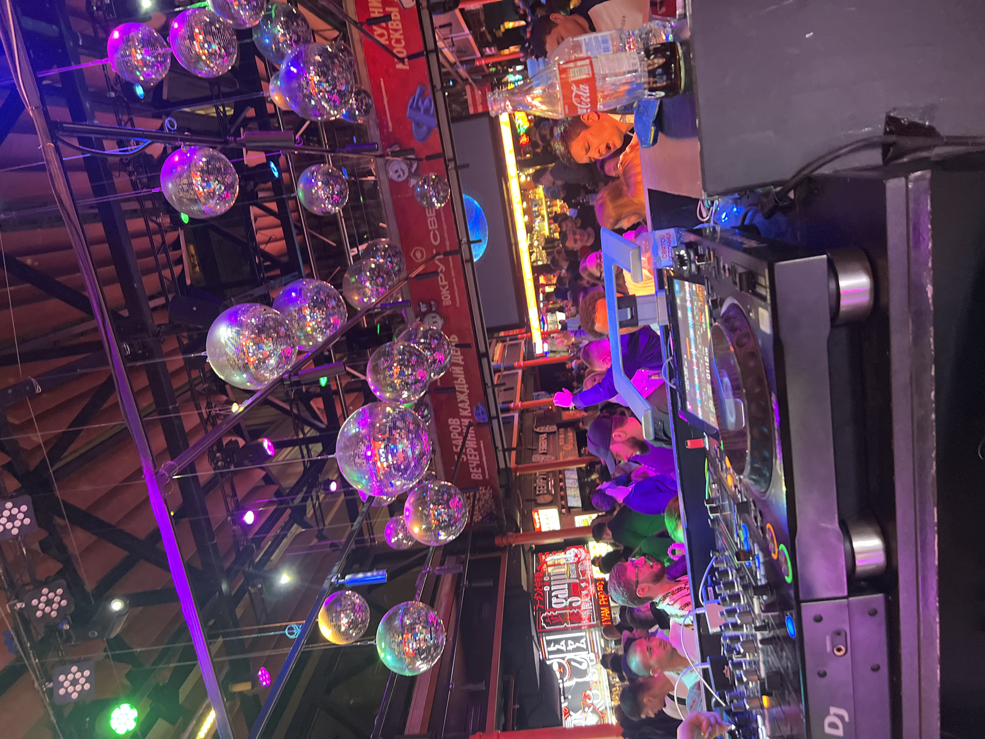 Here is a description of the image:
The image depicts a lively nightlife scene, seemingly inside a club or bar in Moscow, Russia. The focus is on a DJ setup with various controls, dials, and electronic components. In the background, patrons are crowded together, illuminated by colorful lights, contributing to a vibrant atmosphere. Hanging above are numerous disco balls, reflecting the lights and adding to the festive mood. There are also some commercial signs in Russian visible in the background. A Coca-Cola bottle is placed near the DJ equipment.