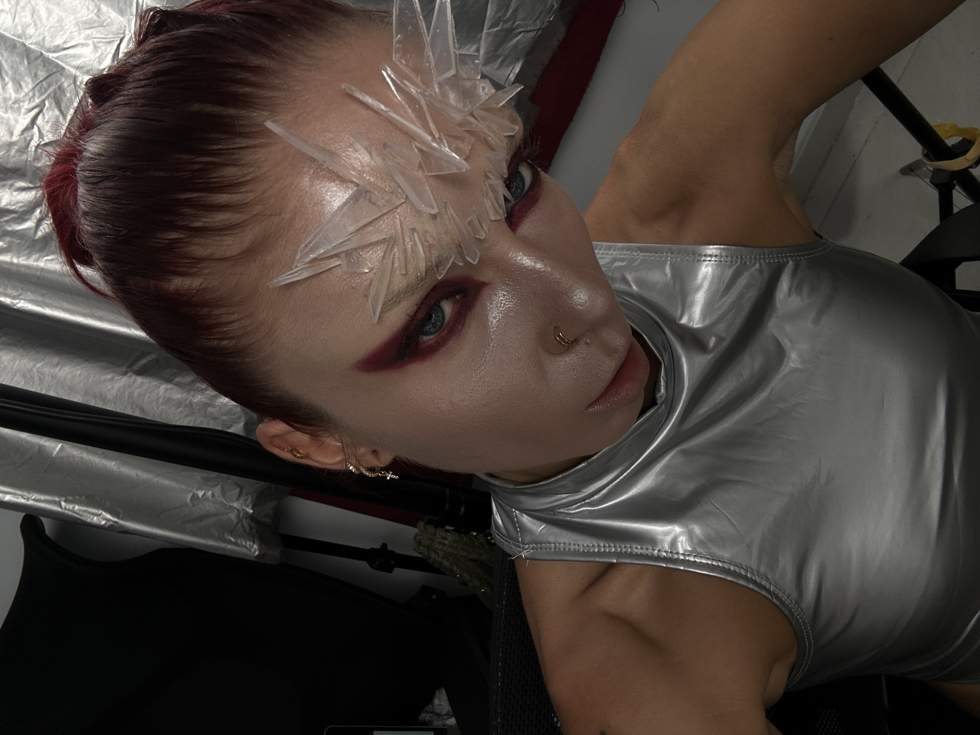 A person with dark red hair is wearing a silver, sleeveless, high-neck top. Their face is tilted down and towards the viewer. They have dramatic makeup, including dark red eyeshadow and what appears to be clear, sharp, jagged adornments on their forehead and cheekbone. The person is wearing gold hoop earrings and a gold nose ring. The background is dark, with elements of silver fabric and some lighting equipment. There are no discernible features indicating a location in North Kuta, Indonesia.