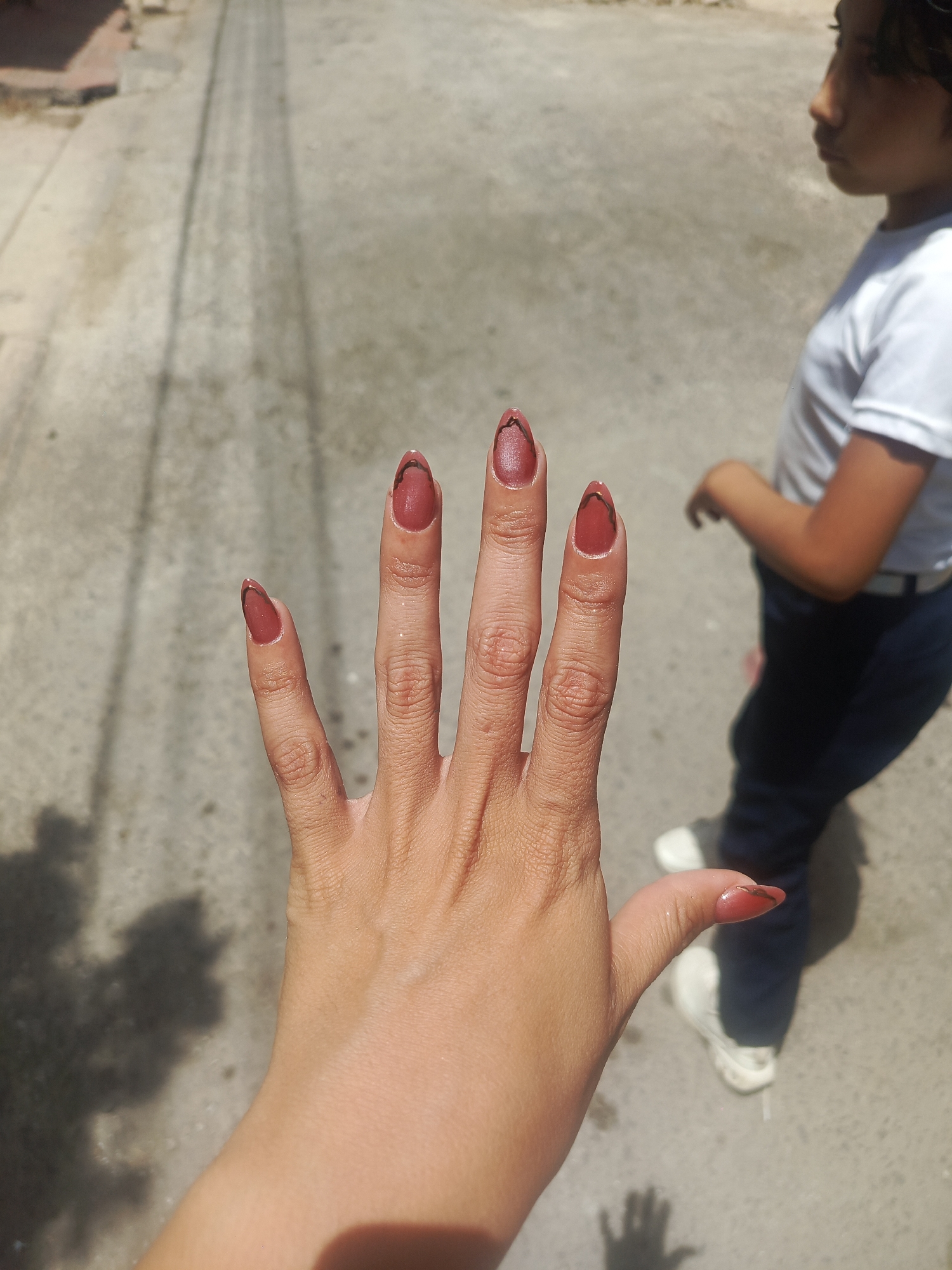 A woman's hand with maroon-colored almond-shaped nails, each featuring a thin black triangle design at the tip, is held up in front of the camera. The background shows an exterior setting, possibly in Juan Griego, Venezuela, with a light gray concrete or asphalt surface and a blurred figure of a child wearing a white t-shirt, blue pants, and white sneakers walking in the background. The lighting suggests a sunny day. The hand casts a dark shadow on the ground.