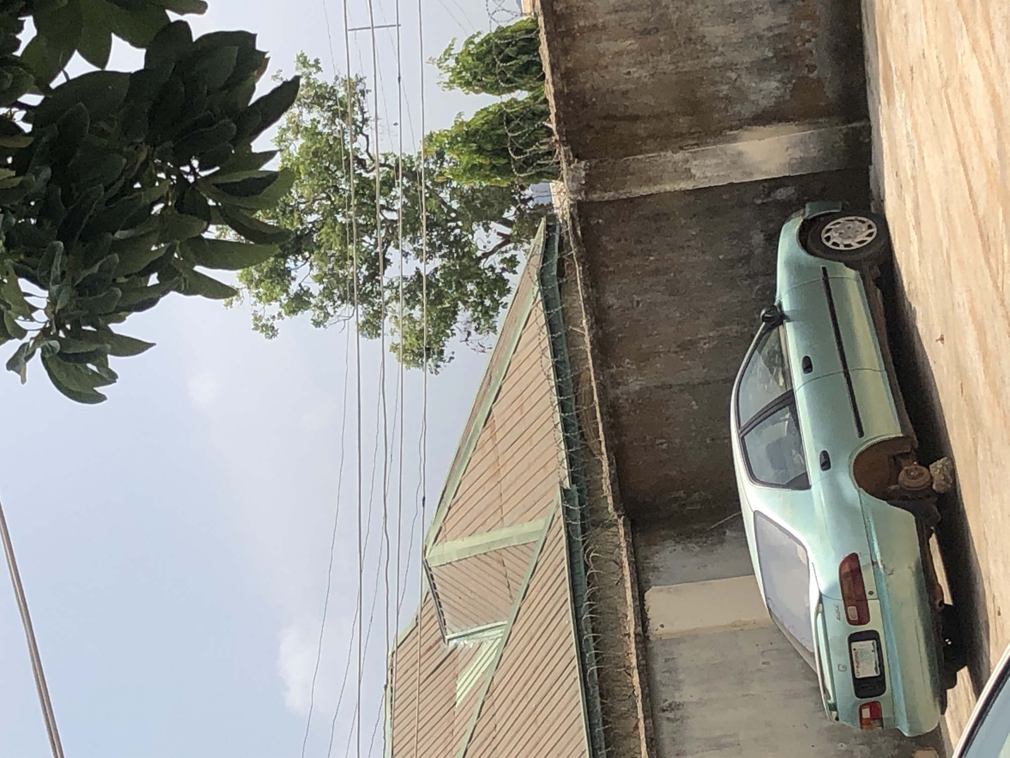 In an outdoor scene in Unknown, Unknown, a light teal sedan with a missing rear wheel sits on a dirt or gravel surface. The car is positioned next to a rough, concrete wall that is topped with barbed wire fencing. Behind the wall, a corrugated metal roof of a building is visible, with a green trim. In the background, a large tree with dense green foliage stands against a pale blue sky with some clouds. The car appears to be abandoned.