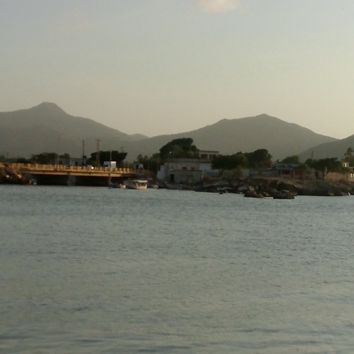A serene waterscape features calm, pale blue water taking up most of the frame. A bridge spans the water with visible supports and yellow guardrails. Beyond the bridge, structures dot the shoreline, interspersed with trees and rocky outcrops. Hazy mountains form the backdrop, their peaks blending into the pale sky. A boat rests near the shore, completing the peaceful scenery.