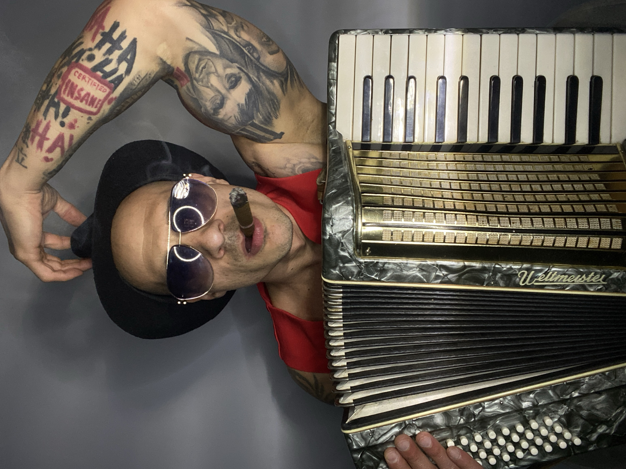 A man is holding an accordion. He is facing the viewer and wearing sunglasses and a black fedora. The man is wearing a red shirt. He has tattoos on both arms. He has a cigar in his mouth. The accordion is a Weltmeister. The background is a gray wall. There is no indication of the location being Pleven, Bulgaria.