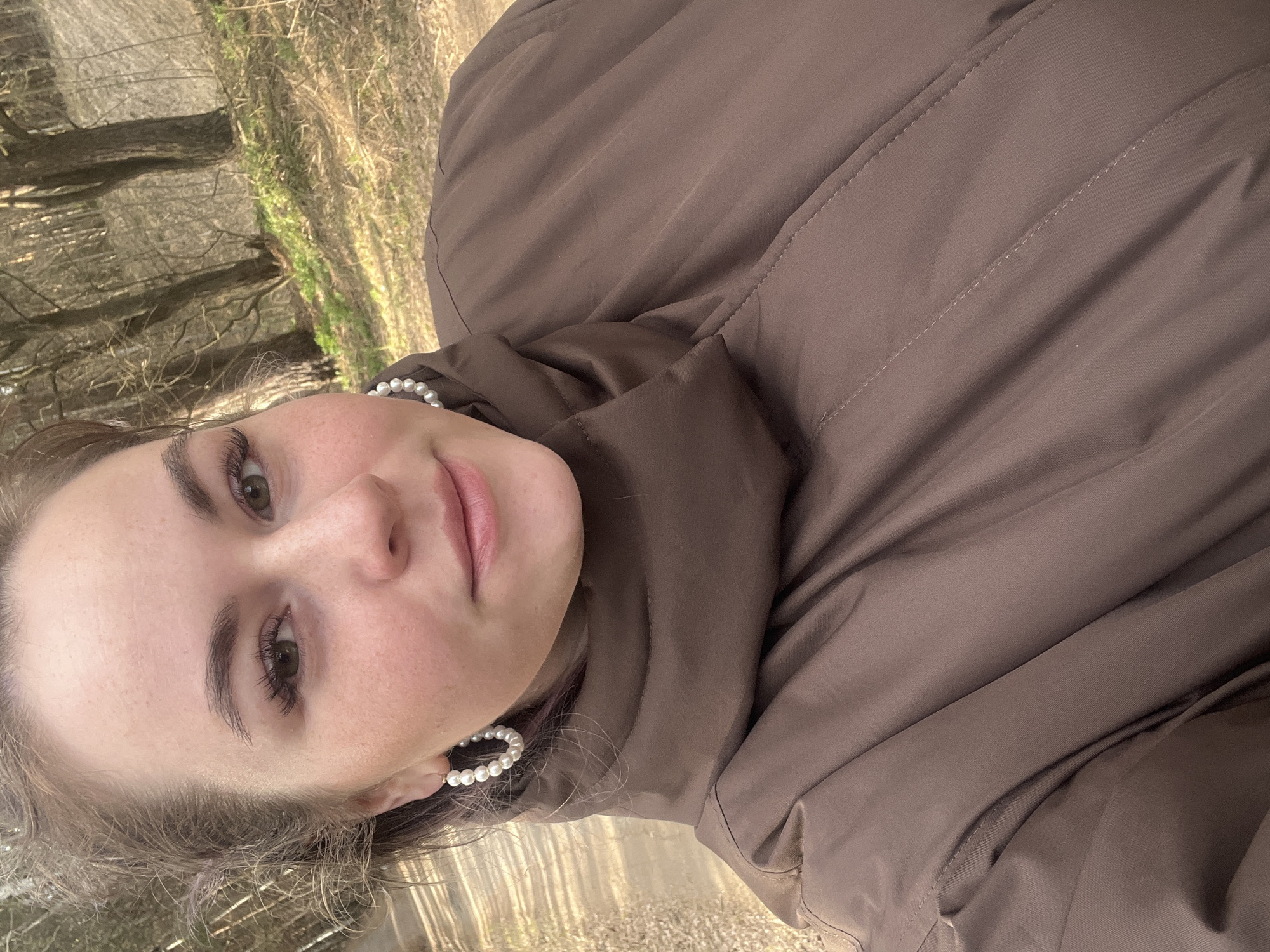 A young woman is captured in a close-up, tilted portrait, appearing to take a selfie or be photographed in an outdoor setting. Her face occupies the left side of the frame, looking directly towards the camera with a soft, neutral expression and slightly parted lips. She has light-colored skin, dark eyebrows, and her eyes are in focus, highlighted by long eyelashes. Her hair is light brown and appears to be pulled back or casually styled, with some strands framing her face.
She is wearing a brown puffer jacket, visible across the right side of the image and covering her shoulders and chest. The jacket has visible stitching patterns and appears warm, suggesting cool weather. Distinctive white pearl hoop earrings adorn her ears, adding a touch of elegance to her casual attire.
The background reveals a wooded or park-like environment, with bare trees and dry, earthy ground cover, indicative of late autumn, winter, or early spring. The natural light suggests it is daytime, with no harsh shadows. The image's diagonal orientation places the natural elements of the background horizontally, contrasting with the vertical subject. No text is visible in the image. Given the location context, this scene is set in Chaikovsky, Russia.