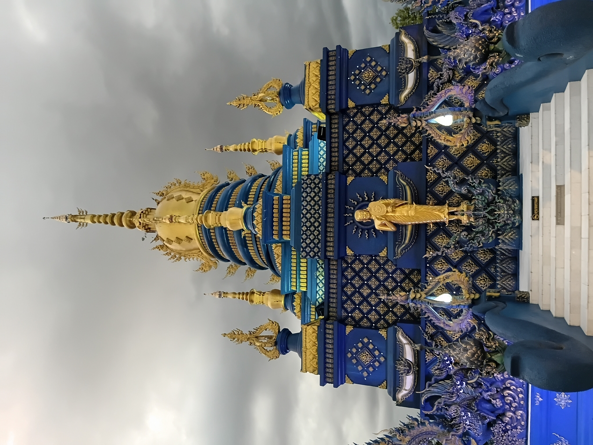 The image shows the Wat Rong Suea Ten, also known as the Blue Temple, in Chiang Rai City Municipality, Thailand. The temple is a vibrant blue structure adorned with intricate gold detailing. In the foreground, a set of white stairs leads up to the temple entrance. Flanking the stairs are ornate blue sculptures of mythical creatures, possibly nagas, and golden statues of Buddhist figures. The temple itself features multiple tiered roofs, each capped with golden spires. A prominent golden statue of a standing Buddha is positioned on the central facade of the temple. The sky above is overcast with dark, dramatic clouds. The image is captured from a low angle, emphasizing the grandeur of the temple.