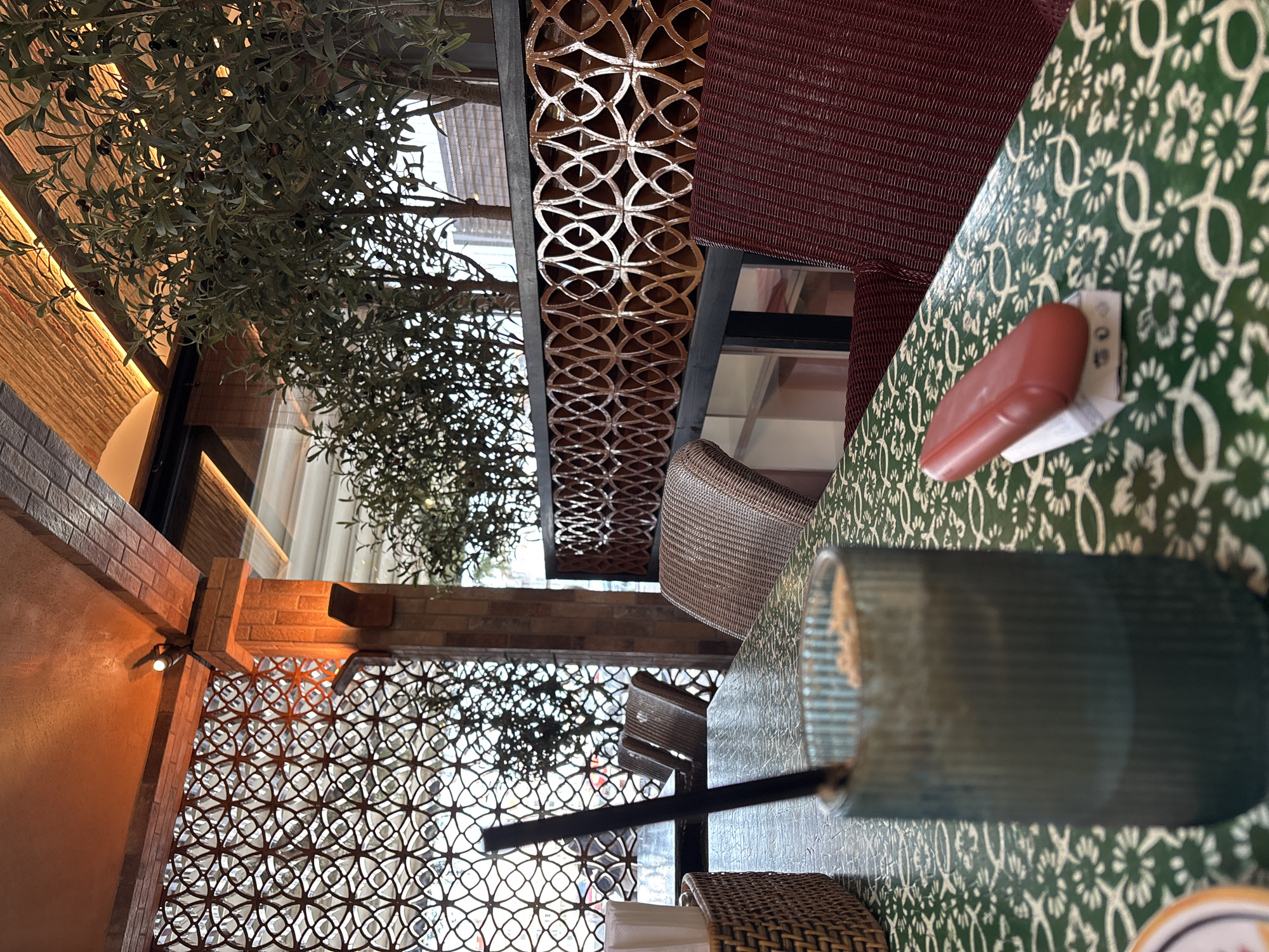The scene is an indoor cafe, likely in Varna, Bulgaria, given the user’s specifications. The focus is on a table setting. The table is covered with a green and white floral patterned surface, and it holds a tall, dark blue glass with a black straw. Beside the glass is a small, red object, possibly a table number or condiment container. A woven chair is partially visible at the table.
In the background, a decorative screen with an interlocking circular pattern provides visual interest. Beyond the screen, glimpses of trees and daylight suggest an outdoor or window-side location. The cafe’s interior also features brick-like wall details and artificial lighting. A maroon-colored woven seat covering is also partially visible.