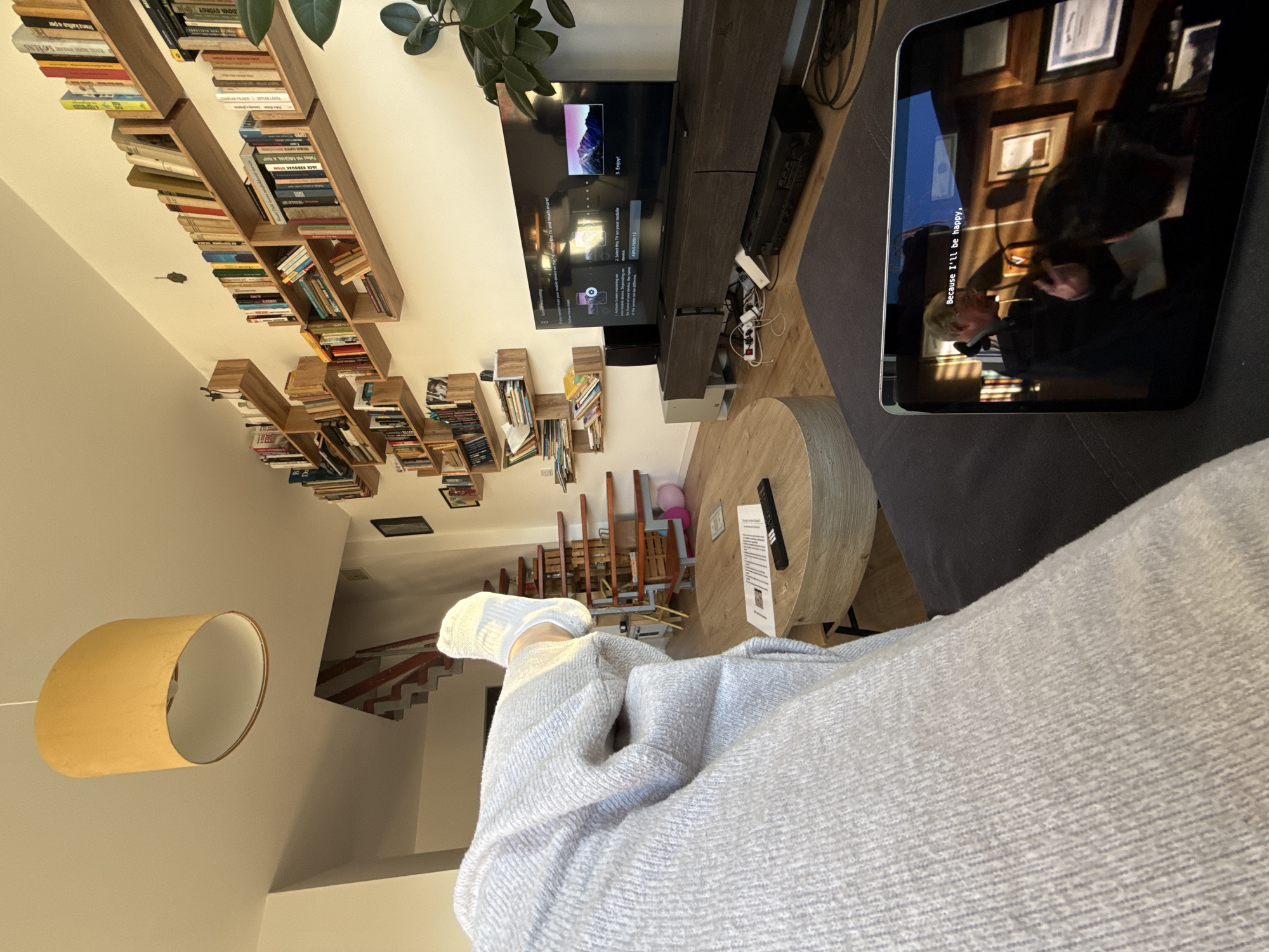 This image captures a relaxed indoor scene, likely a living room, from a first-person perspective, looking over a person's legs towards a television and extensive bookshelves. The viewer is positioned as if lounging on a sofa, with their legs, covered by a light grey, textured blanket or robe, and feet in white socks, extended into the frame.
To the right, a tablet displays video content with the visible subtitle, "Because I'll be happy,". A round wooden coffee table is positioned in front of the sofa, holding a black remote control and some papers with illegible text.
In the mid-ground, a large flat-screen television is mounted above a dark wooden media console. The TV screen shows an interface for connecting mobile devices, displaying text like "Select this TV on your mobile," "43PUS7609/12" (a model number), and "Enjoy!". The word "Depending" is also partially visible. Clutter of cables and electronic devices can be seen on and around the media console.
The background features a striking arrangement of modular wooden bookshelves, filled with numerous books, some of which are partially legible, including "DOVA GYÖRGY," "SPAENS," and "franz kafka a per." A large green plant is visible in the upper left corner. A beige cylindrical pendant lamp hangs from the ceiling to the lower left, and a wooden staircase is visible in the distance, indicating a multi-level home.
The scene depicts a peaceful daytime moment of leisure in a contemporary home in Budapest, Hungary, with the person enjoying media content.