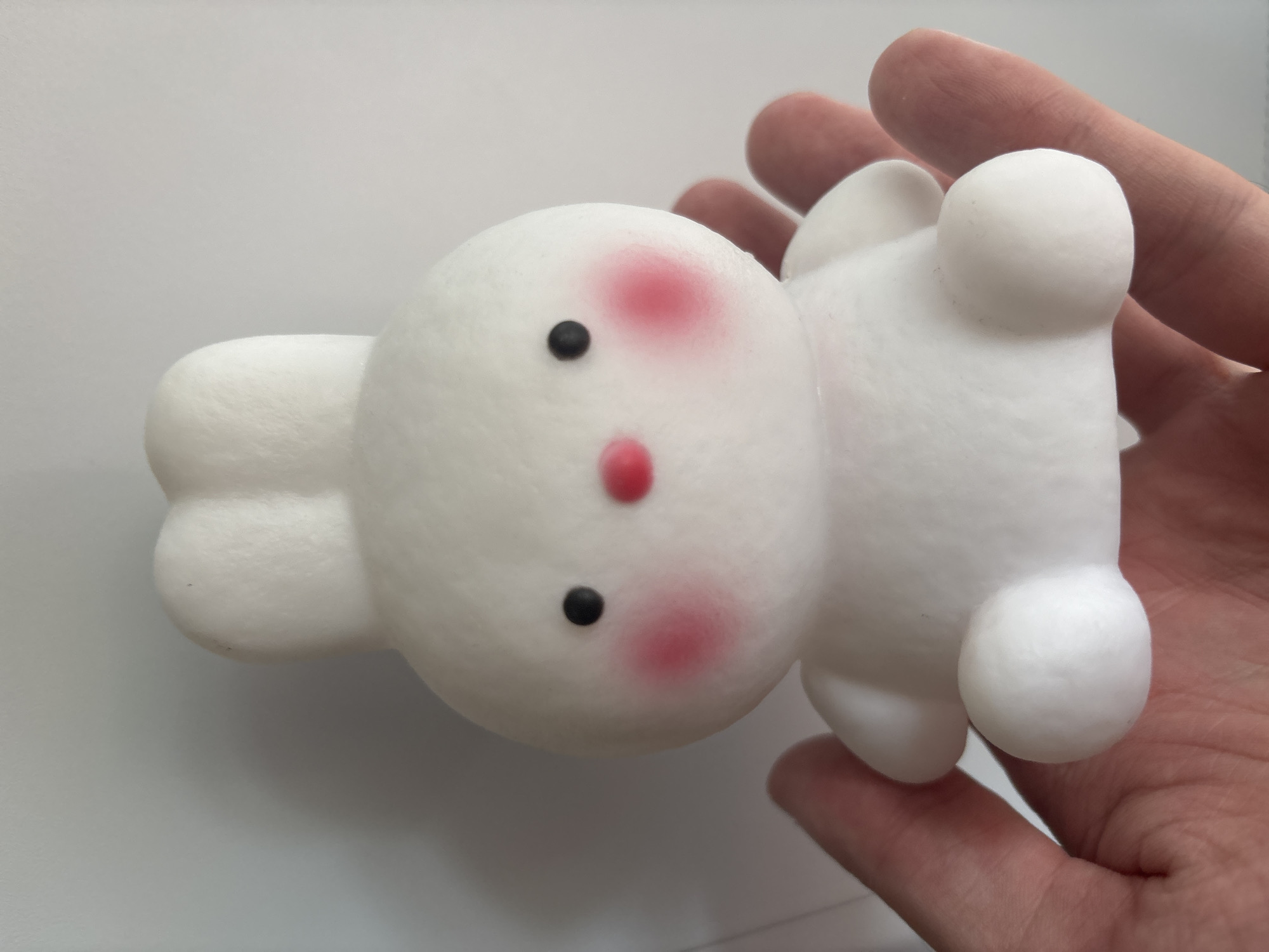A hand is holding a white, squishy toy that resembles a round, cartoonish character. The toy has a simple face with two black dots for eyes and a red oval for a nose. Pink blush marks are on its cheeks. The toy is lying on its back, with its rounded limbs extended outwards. The hand supporting it is a light skin tone, with visible fingers and palm. The background is a plain, light gray surface. The lighting appears to be even and diffused, suggesting an indoor setting with artificial light or an overcast day. There is no visible text in the image. The overall impression is that of a playful, soft toy being held.
