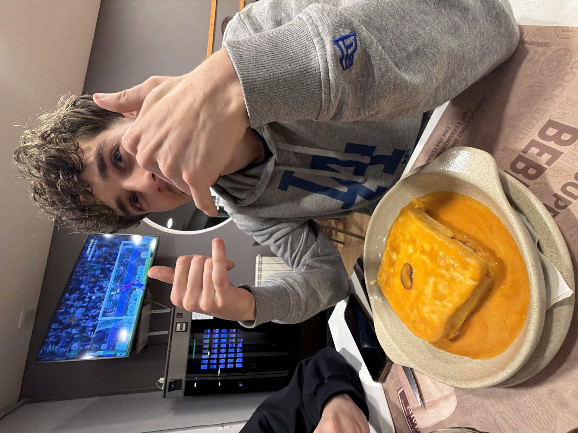 Here's a description of the image:
A young man in a gray hoodie with a "LA" logo is seated at a table with a plate of Francesinha, a Portuguese sandwich, in front of him. He's gesturing towards the food with his hands. In the background, a TV screen displays a soccer match, and there's a payment kiosk nearby. The table has a printed paper placemat, and the focus is on the man and the food, likely in a restaurant or cafe. The location is Bragança, Portugal, based on the food being served.