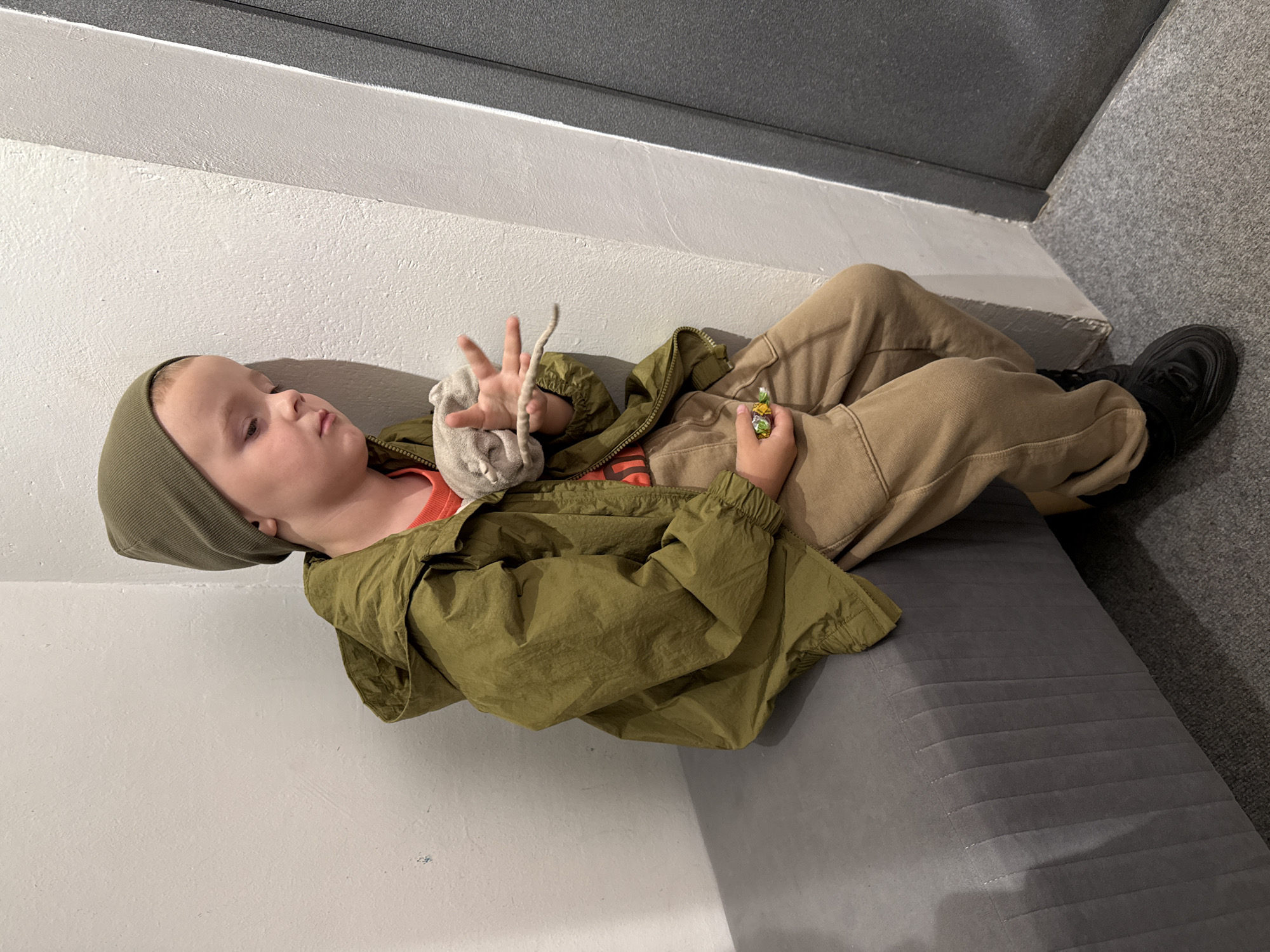 A young Caucasian boy, appearing to be around 4-6 years old, sits on a gray upholstered bench, leaning his head against a white textured wall. He is wearing a green beanie, a green hooded jacket zipped over an orange t-shirt, and tan sweatpants. His left hand holds a small, wrapped candy, and his right hand is extended, holding a light gray stuffed toy that resembles a mouse with a long tail. The boy looks down at the toy, appearing somewhat melancholic or contemplative. The background consists of a white wall and a dark gray door or partition. The floor is covered with gray carpet. The scene is well-lit, suggesting an indoor environment. The overall impression is of a child in a moment of quiet reflection or perhaps mild disappointment. No specific identifying landmarks or context to place this image in Chaikovsky, Russia are visible.
